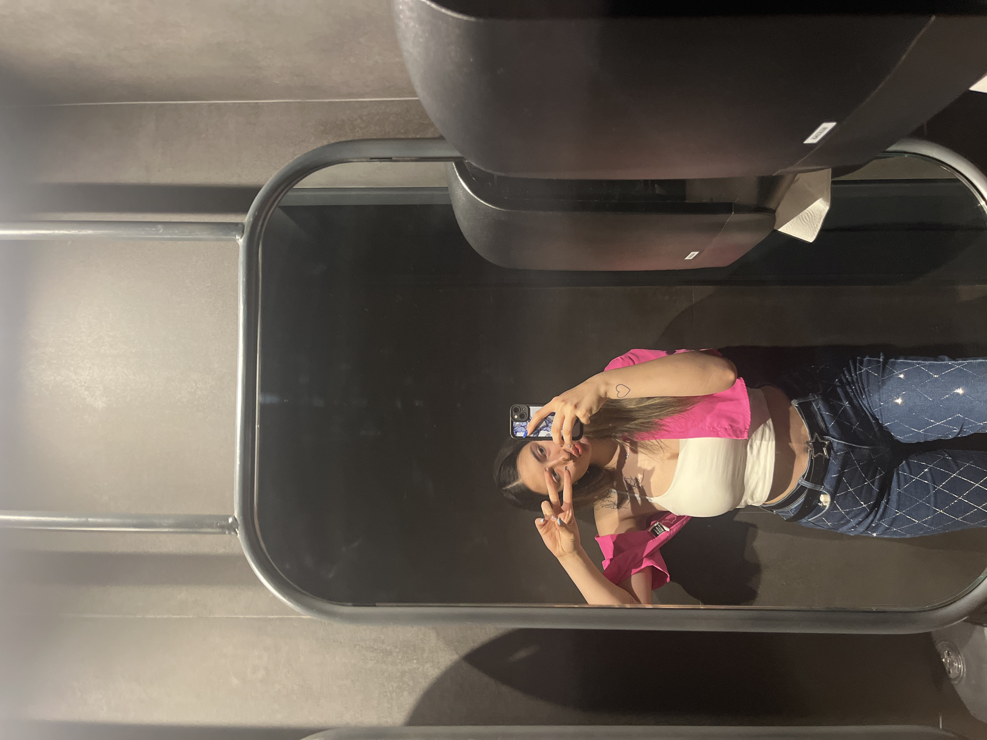 A young woman with long, dark hair is taking a selfie in a public restroom. She is wearing a white tank top and blue jeans with a diamond pattern. She has a pink jacket draped over her shoulders and is holding her phone with her right hand, making a peace sign with her left hand. A heart tattoo is visible on her left arm. The background shows a textured gray wall, a large mirror, and what appear to be two paper towel dispensers. The location is the restroom of a establishment in Plovdiv, Bulgaria.