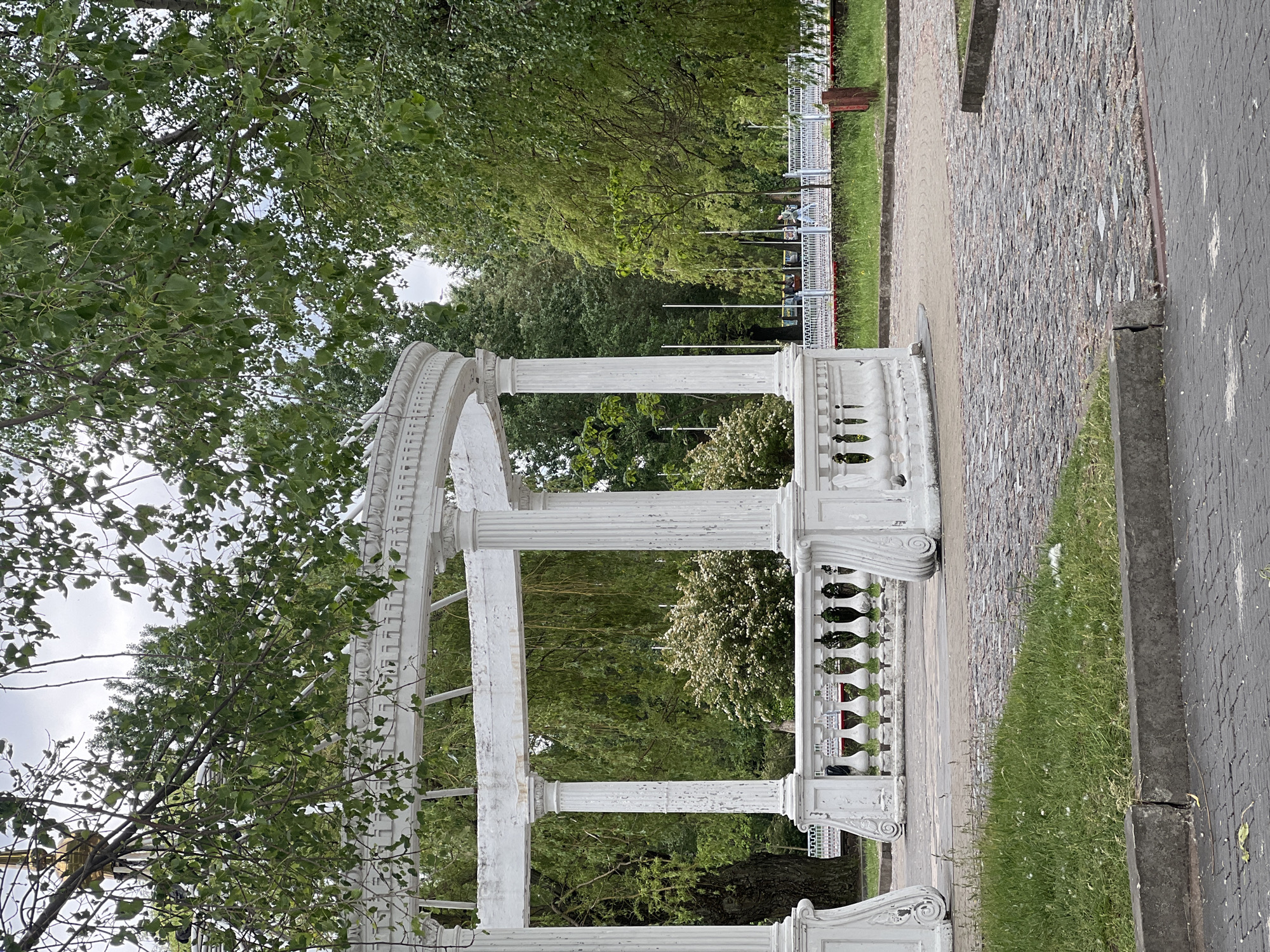 The image depicts a weathered, white pergola situated in a park. The pergola features classical architectural elements, including columns and balustrades, showing signs of aging with peeling paint. The structure is partly surrounded by lush greenery, including trees with dense foliage. The ground is a mix of paving stones, concrete, and grass. In the background, a white fence runs along the edge of the park.
