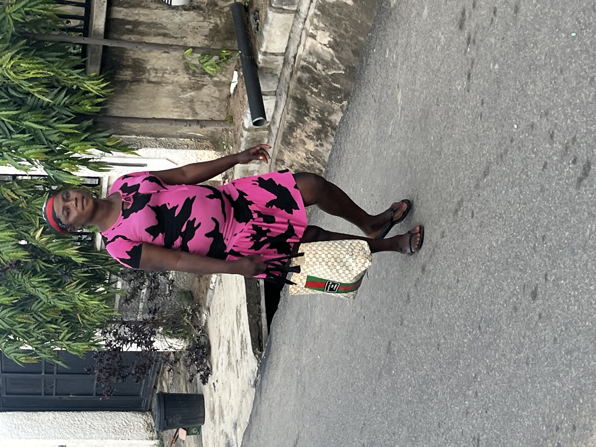 An individual walks on an asphalt road in Abuja, Nigeria. The person is wearing a pink dress with a black abstract pattern and a red headscarf. They are carrying a patterned tote bag with green and red stripes and a logo. The environment includes a textured white wall on the left, a black metal gate, a trash bin, lush green trees and plants, and a concrete barrier with a black pipe. The road surface is grey and shows signs of wear.