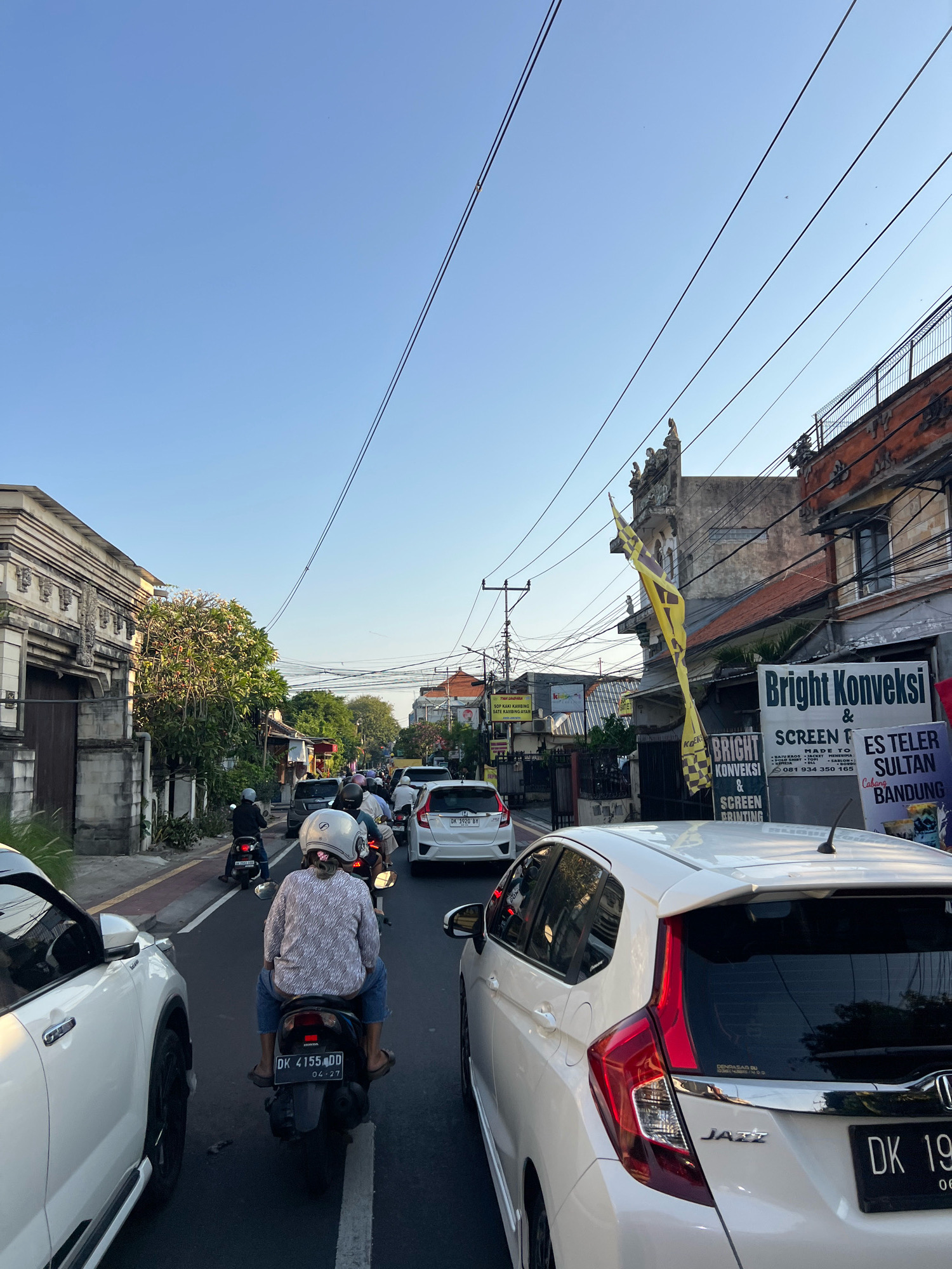 The image depicts a congested street scene in Denpasar, Indonesia, likely during late afternoon given the low angle of the sun casting long shadows. The foreground is dominated by the rear of a white Honda Jazz, its taillights illuminated. To the left, another white SUV is partially visible. The road is filled with a mix of motorcycles and cars, creating a traffic jam.
Several individuals are on motorcycles, most wearing helmets. One rider in the middle foreground is a woman wearing a patterned shirt and jeans, her motorcycle displaying a license plate starting with "DK 4155 DD". Another motorcycle rider further back has a white helmet and is wearing a grey and white patterned shirt.
Buildings line both sides of the street. On the left, a traditional Balinese-style building with intricate stone carvings is visible. On the right, several businesses are apparent. One sign reads "Bright Konveksi & SCREEN PRINTING" with a phone number listed. Another sign, further down the street, indicates "ES TELER SULTAN BANDUNG". Yellow and black checkered flags are also displayed, possibly related to a business or event. Power lines crisscross overhead, typical of urban environments. The sky is clear blue with a few distant birds visible. The overall atmosphere is one of a busy, everyday urban commute in Bali.