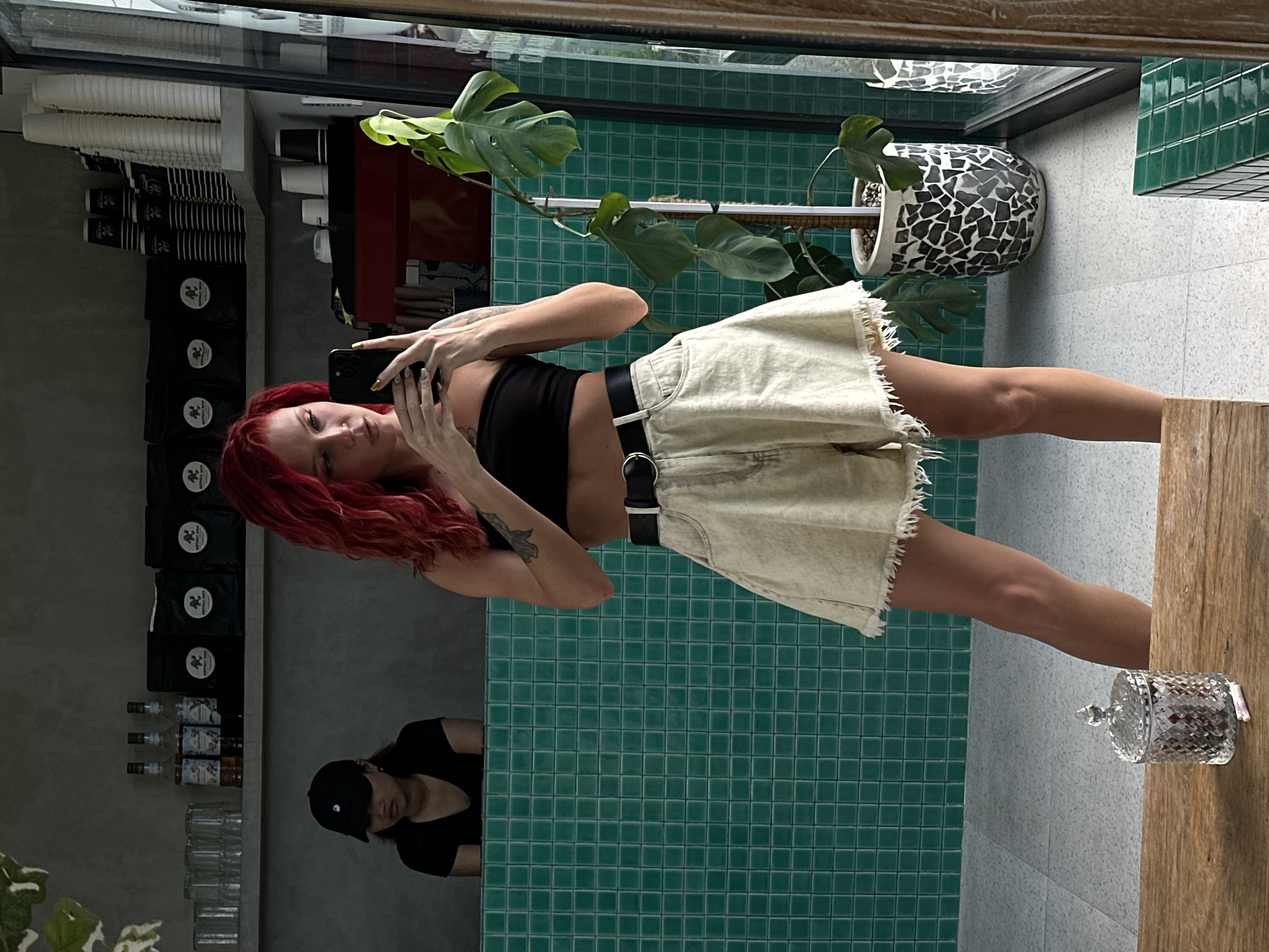 A woman with long red hair wearing a black crop top and a light tan mini skirt with a black belt stands in front of a reflective surface in a cafe in North Kuta, Indonesia. She is holding a black smartphone and taking a photo of herself. Behind her, the background shows a display of coffee beans, a coffee machine, and a person wearing a black cap. A potted monstera plant sits on a shelf above the woman. To her right, a glass container sits on a wooden surface. The cafe features green square tiles on the wall behind the woman and white tiled walls adjacent to the wooden surface.