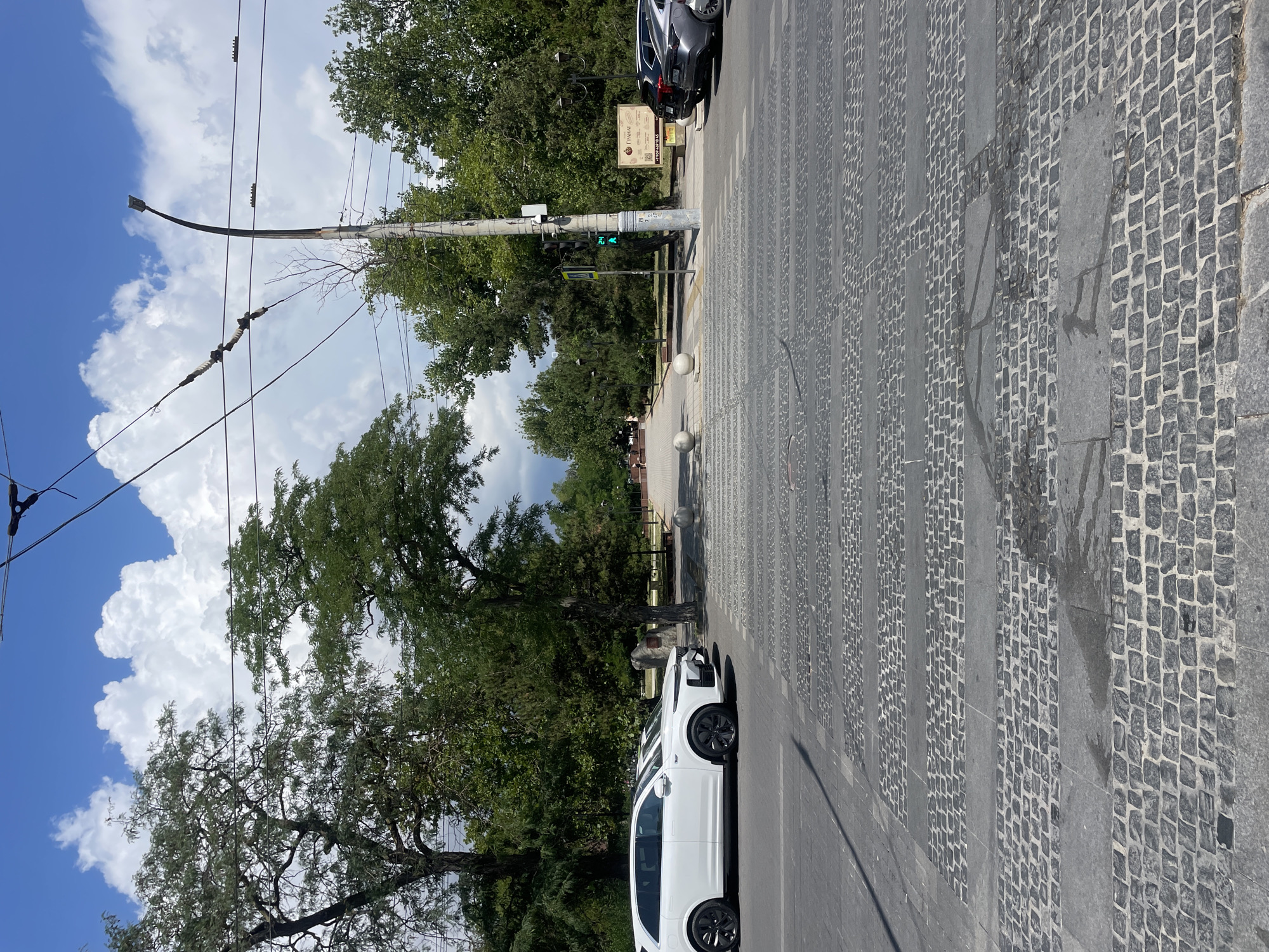 The image depicts a pedestrian crossing with a distinct cobblestone pattern on a street in Novorossiysk, Russia. A white SUV is parked partially on the crossing, and another car is visible behind it. The background features lush green trees and a cloudy sky, typical of a summer day. A traffic light is visible, indicating an intersection. In the distance, people can be seen walking in a park-like area, suggesting a public space. The overall impression is of a bustling urban environment with a touch of natural scenery.