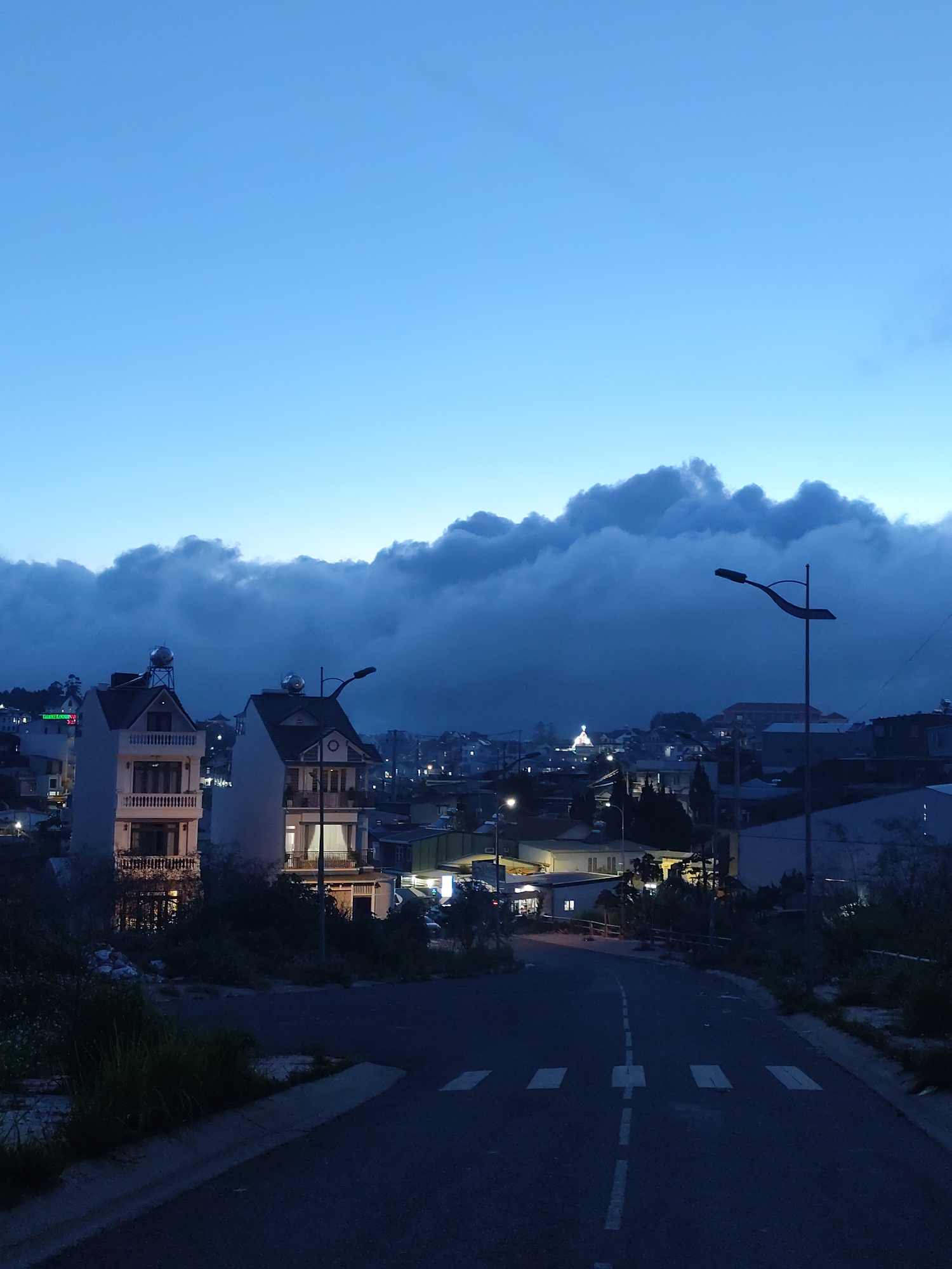The image shows a street scene at dusk or dawn in Lang Biang - Đà Lạt, Vietnam. The sky is a gradient of blue, with a thick layer of clouds obscuring the horizon. In the midground, two tall buildings with balconies and water tanks on their roofs are visible. Streetlights illuminate the road and surrounding houses. A road with pedestrian crossings and a dashed center line leads towards the city. The foreground shows a portion of the road and roadside vegetation. The overall lighting is dim, with visible artificial lights from buildings and streetlamps. A church or other notable building with illuminated spires can be seen in the distance.