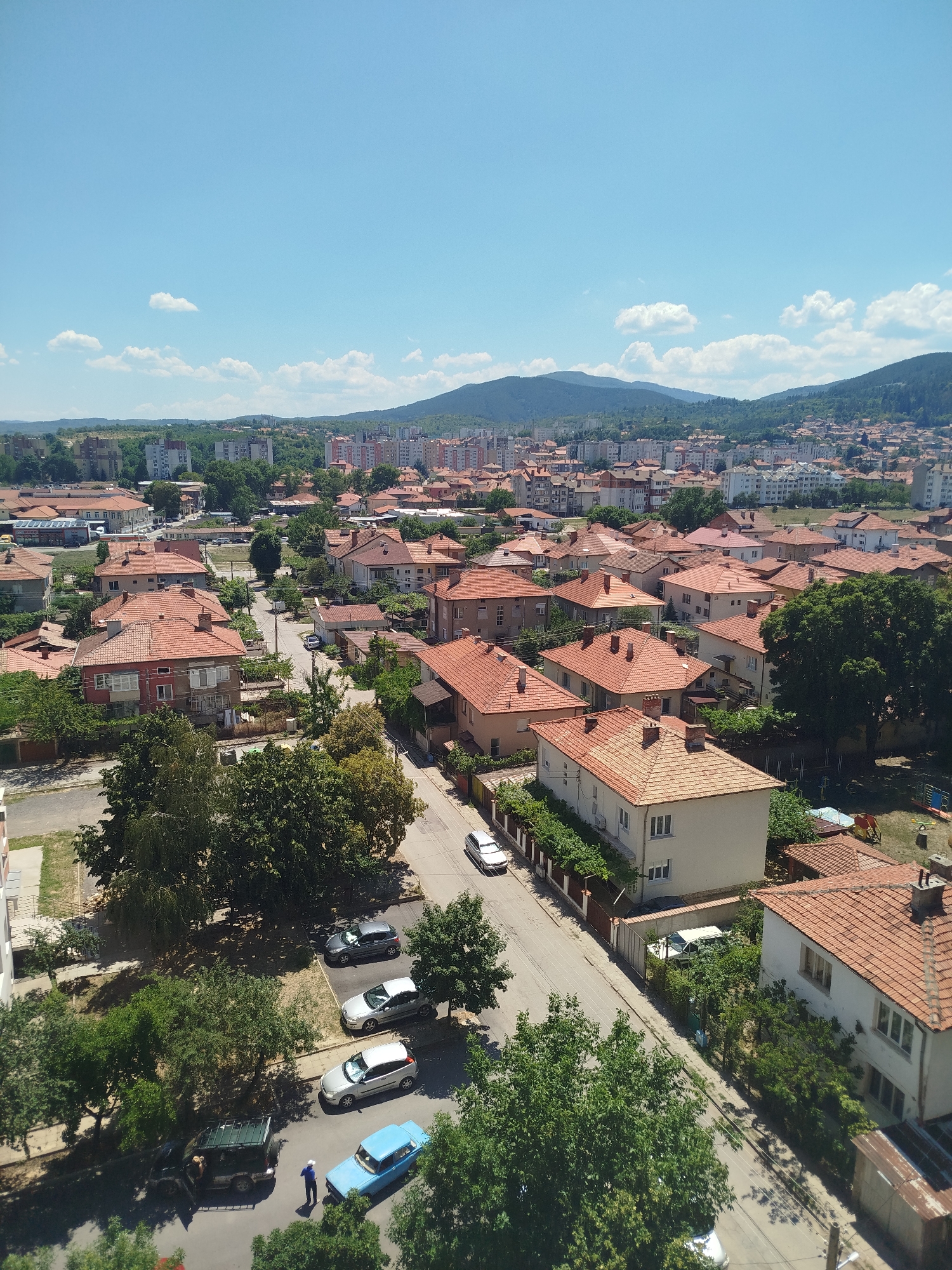 An aerial view captures a residential area in Kyustendil, Bulgaria, under a clear blue sky with scattered clouds. The scene is dominated by numerous houses with characteristic red-tiled roofs, interspersed with trees and vegetation. A road winds through the neighborhood, with several parked cars visible. In the foreground, a few houses are prominently featured, showcasing their architecture. Further in the distance, a denser urban landscape with taller buildings emerges, leading to rolling hills and mountains in the background. The overall impression is of a sunny day in a European town, with a mix of older and more modern structures.