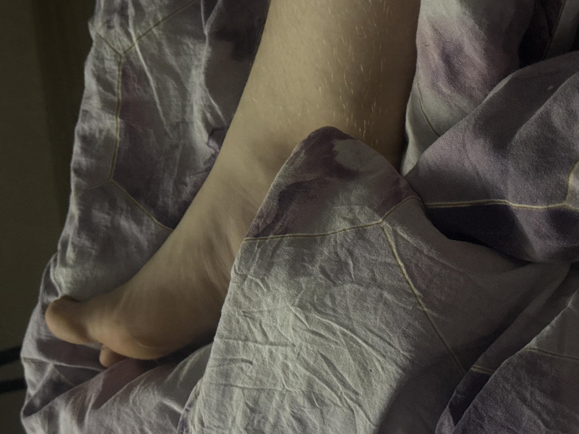 This media file shows a close-up view of a person's bare leg and foot covered by a purple and white patterned comforter. The comforter has creases and folds, suggesting it is in use, possibly on a bed. The leg is lightly covered in fine hairs, and the skin tone is fair. The lighting suggests a dimly lit environment, possibly indoors during the evening or night, or in a room with closed curtains. The scene is intimate and personal, focusing on a small detail of a person resting. There are no visible text elements in the image. The provided location context of Saratov, Russia, does not offer specific cues visible within the image itself.
