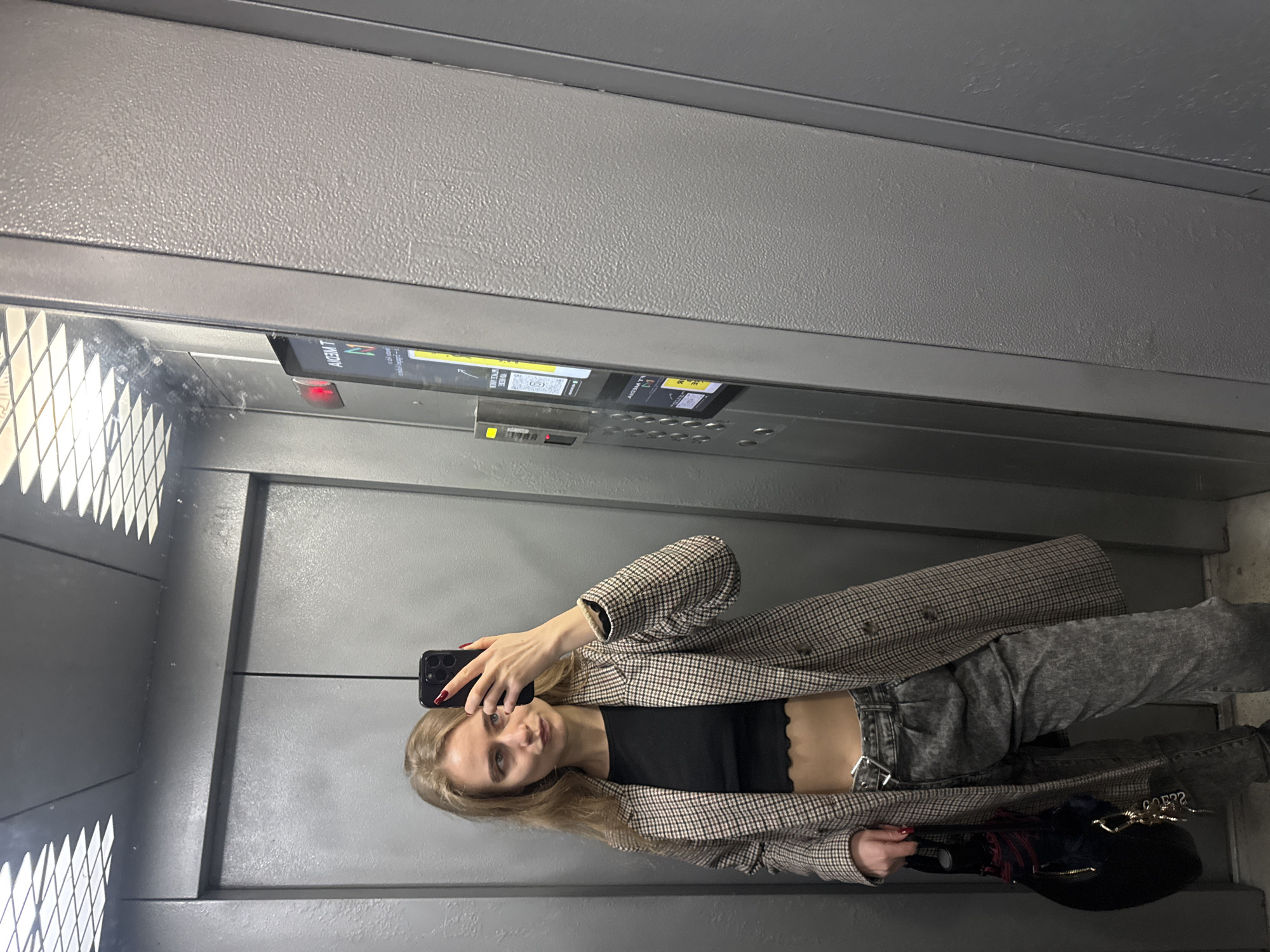 A woman is taking a selfie in an elevator. The elevator has a metal interior with a display panel. The woman is wearing a plaid blazer, a black top, and grey jeans. She is holding a black handbag. The location appears to be indoors in the city of Unknown, Unknown.