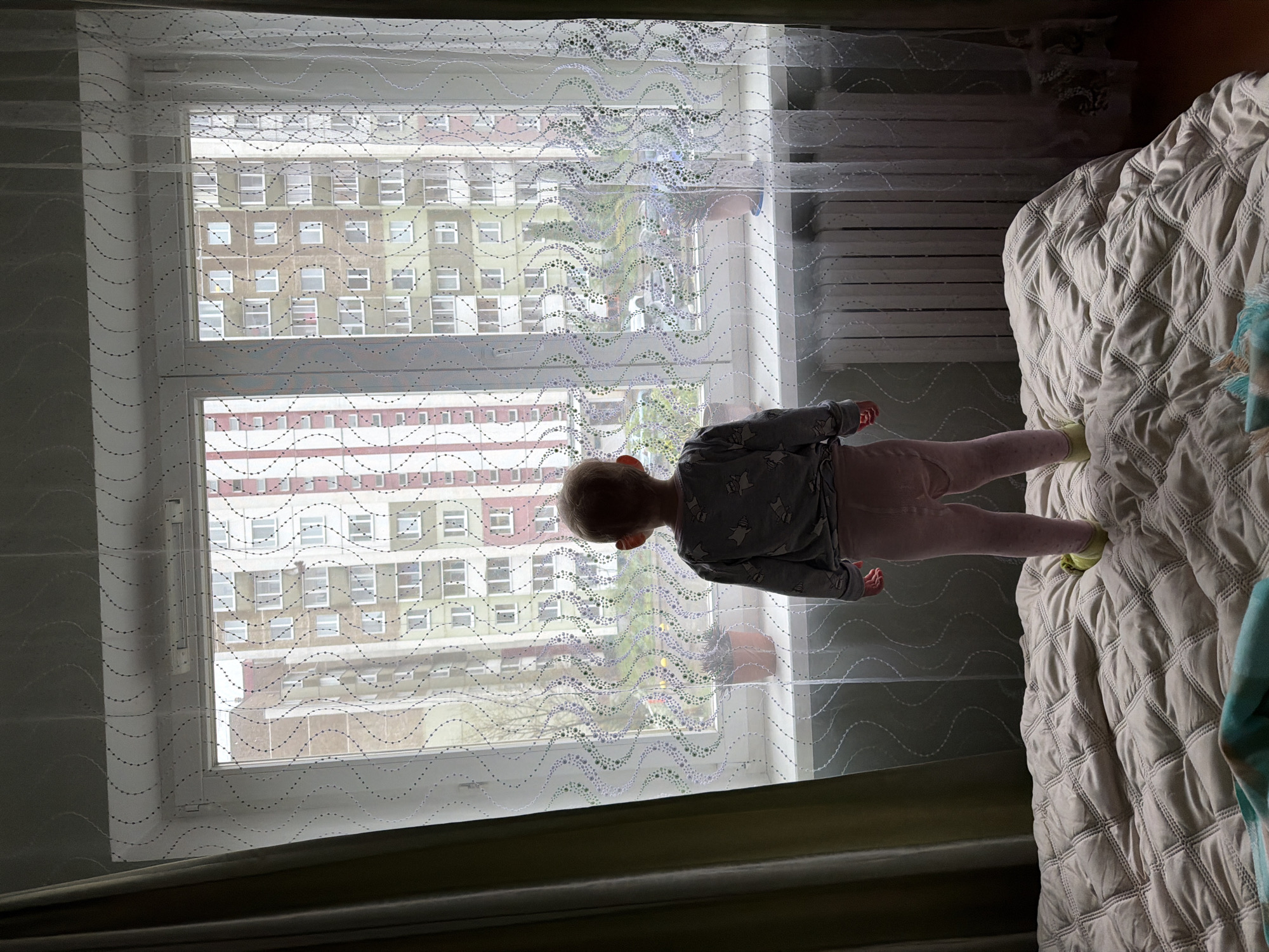 A child stands on a bed near a window, looking outside. The window is covered with a semi-transparent curtain with a wavy dotted pattern. Outside the window are buildings with multiple floors and windows. The child wears a gray shirt with a pattern and pink pants. The bed has a white quilted cover. The environment suggests an indoor setting. It's not possible to define a location within a city based on the image alone.