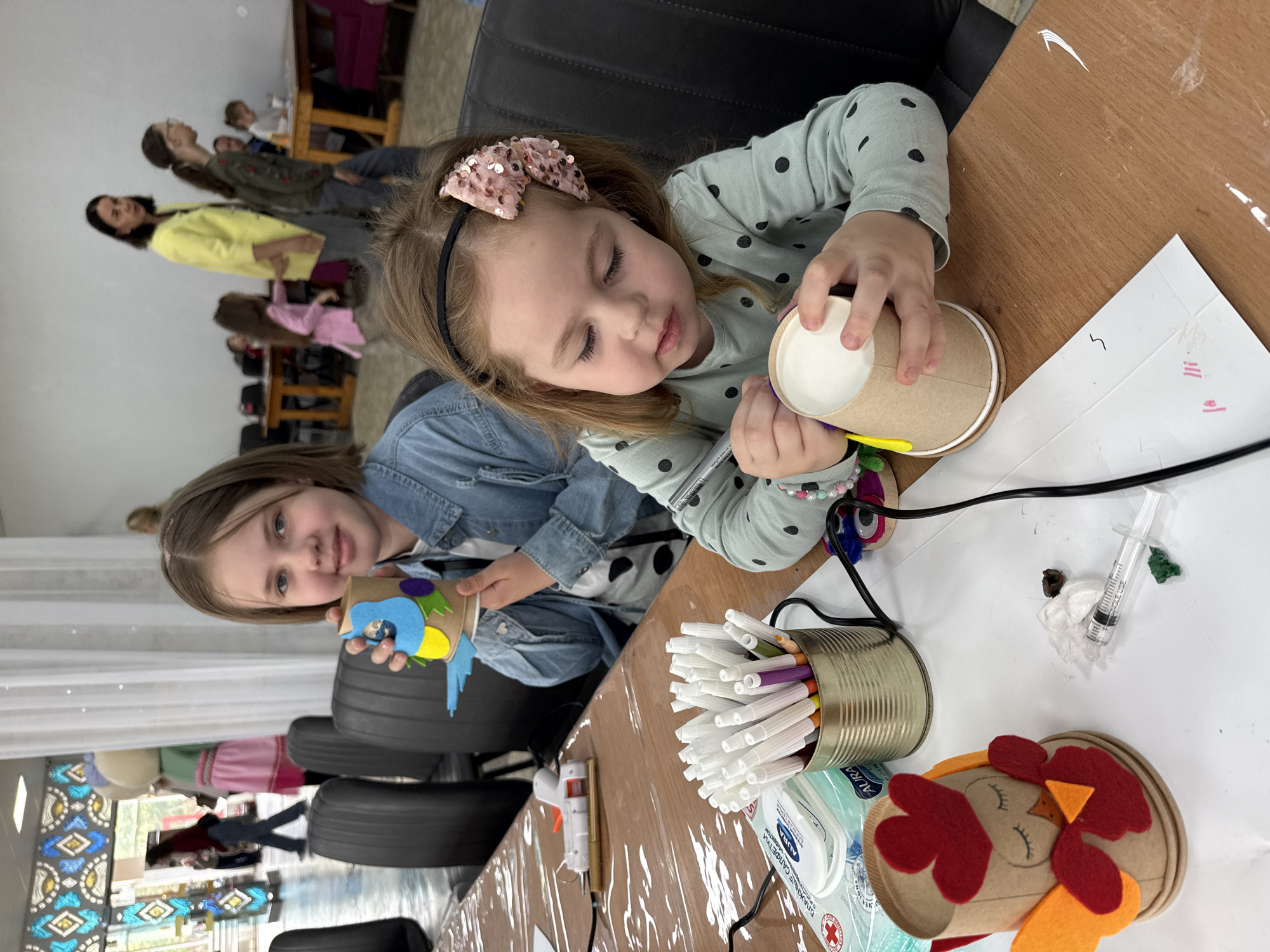 Two young girls are seated at a table in what appears to be a classroom or community center in Izhevsk, Russia, based on the logo on a package of wet wipes in the lower foreground. The girls are engaged in an arts and crafts activity, decorating cups. The girl on the right, with a bow in her hair, is examining a cup, while the girl on the left holds up a decorated cup. Various craft supplies, including markers, a glue gun, and felt shapes, are scattered on the table. In the background, other people are seated at tables, suggesting a group activity.