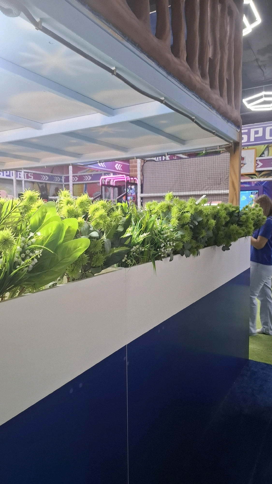 The image depicts an indoor entertainment venue, likely an arcade or a children's play area, with a distinct tropical or jungle theme. In the foreground, a white planter box is filled with lush, artificial greenery, featuring large, broad green leaves interspersed with spiky green seed-like pods and delicate white flowers resembling lily of the valley. This planter box runs horizontally across the lower half of the frame.
Behind the planter, a multi-tiered structure with a brown, bark-like texture forms an overhead walkway or barrier, evoking tree trunks. This structure is supported by pale blue panels and beams, suggesting an elevated platform or ceiling. Cables are neatly secured along the underside of these blue panels.
In the background, a variety of arcade games and attractions are visible, illuminated by neon pink and purple lights. Signage with white text and decorative arrows is present, and a netted enclosure suggests a climbing or play area. The overall impression is of a vibrant, engaging space designed for recreation.
To the right of the image, a woman wearing a dark blue t-shirt and light-colored pants stands, facing away from the camera and towards the arcade games. She appears to be observing the attractions or interacting with the environment. The floor in the foreground transitions from a dark blue carpet to a lighter blue and then white angled sections. The scene is brightly lit, consistent with an indoor commercial establishment.
This venue is located within the MEGA Park in Shymkent, Kazakhstan, a popular shopping and entertainment complex.