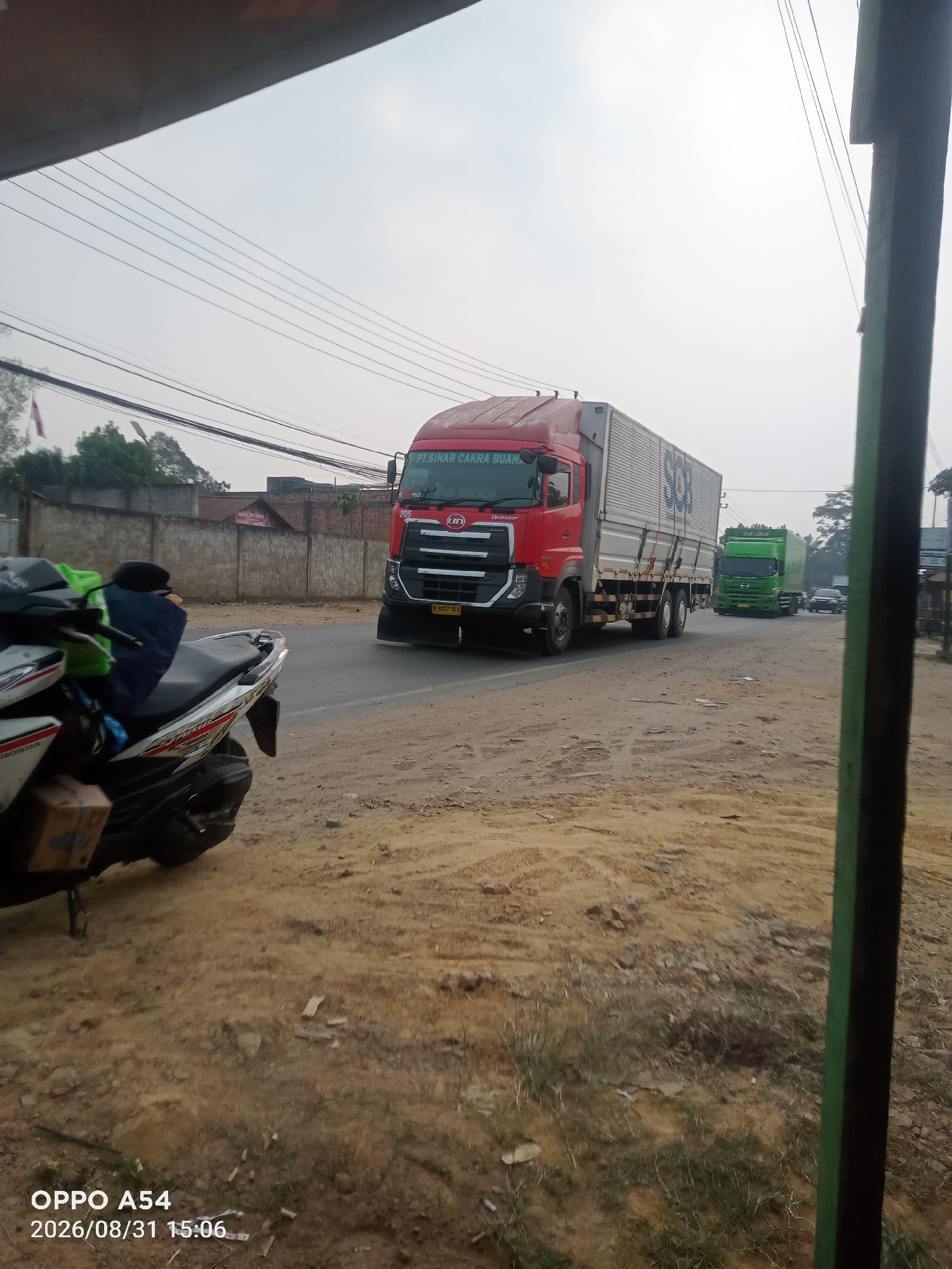 A side-view, eye-level, medium shot captures a street scene in Jambi, Indonesia. A red semi-trailer truck with a white cargo container is on the right side of the road, moving away from the viewer. The truck has "PT SINAR CAKRA BUANA" and "Quester" visible on its exterior. To the left of this truck, a green semi-trailer truck is also visible on the road, further down the street. In the foreground on the left, a parked motorcycle is partially visible. The road surface is dirt and gravel. A concrete wall runs along the left side of the road, with some buildings and trees visible beyond it. Power lines crisscross the sky. The sky is overcast and appears to be daytime. A timestamp in the bottom left corner reads "OPPO A54 2026/08/31 15:06".