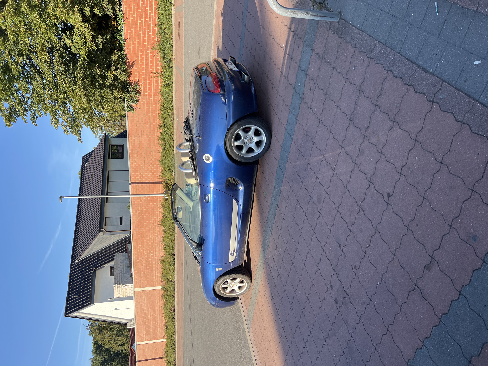 A blue convertible sports car is parked on a red brick paver walkway. The car is facing left and is viewed from the side. The car has its top down, revealing tan leather seats and chrome roll bars. There are two exhaust pipes visible at the rear of the car. The walkway is next to a paved road. Behind the walkway is a low, red brick wall, topped with green bushes. Beyond the wall are houses and trees. The sky is clear and blue, with a faint white contrail visible. The lighting suggests it is daytime, likely morning or afternoon, given the long shadows cast by the car and the sun's angle.