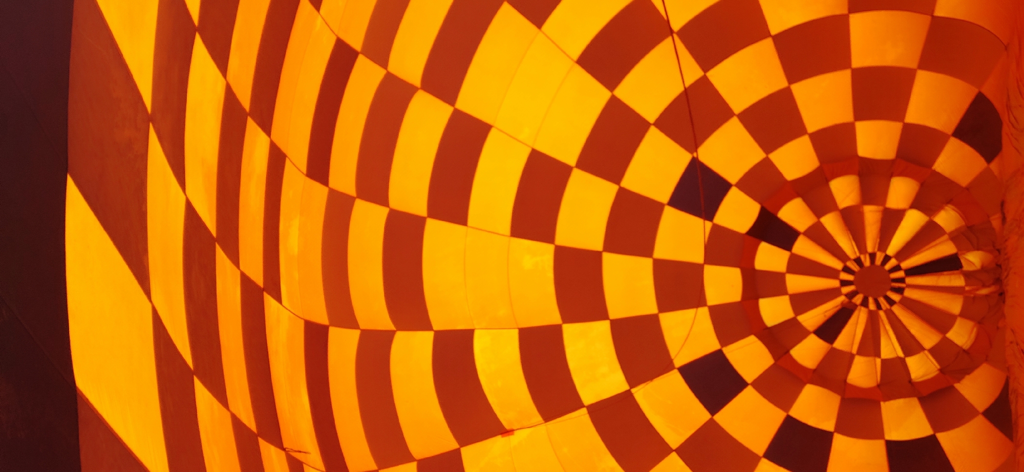 The image depicts the interior of an orange and brown checkered hot air balloon. The pattern is a grid of squares, which appear warped and curved due to the balloon's spherical shape. The perspective is from within the balloon, looking upwards towards the opening, which is a circular void with radiating segments. The lighting suggests the balloon is illuminated from the outside. There are no people or specific environmental features of Kalath, India visible in this image.