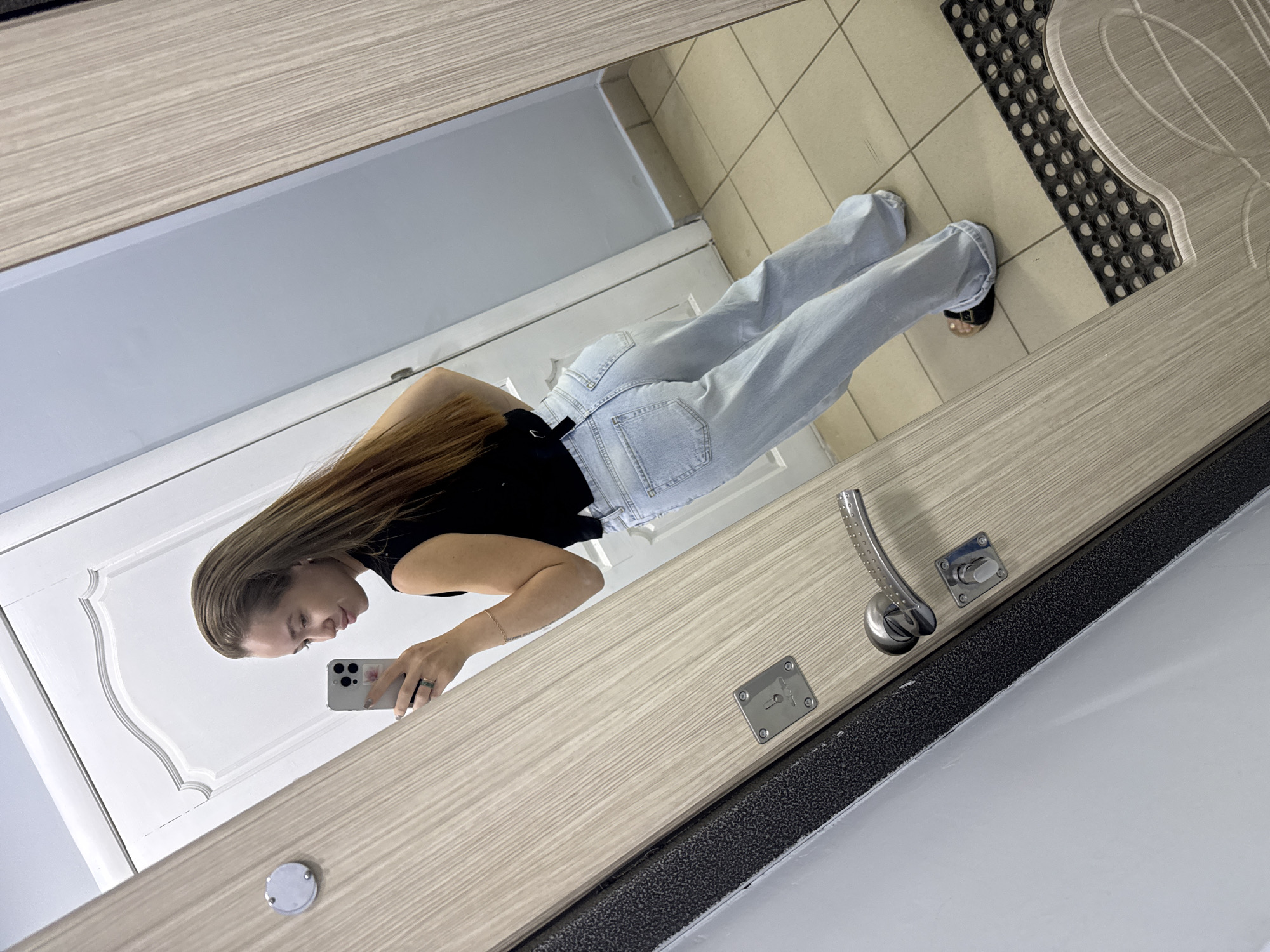 A woman stands in front of a mirror in Chaikovsky, Russia, taking a selfie with a smartphone. She is wearing a black top and light blue jeans. Her long brown hair is down. The mirror reflects a tiled floor, a white door, and a gray wall. The door frame has a lock and handle. A black and gray rug is visible near the door. The mirror is mounted on a door in Chaikovsky, Russia.