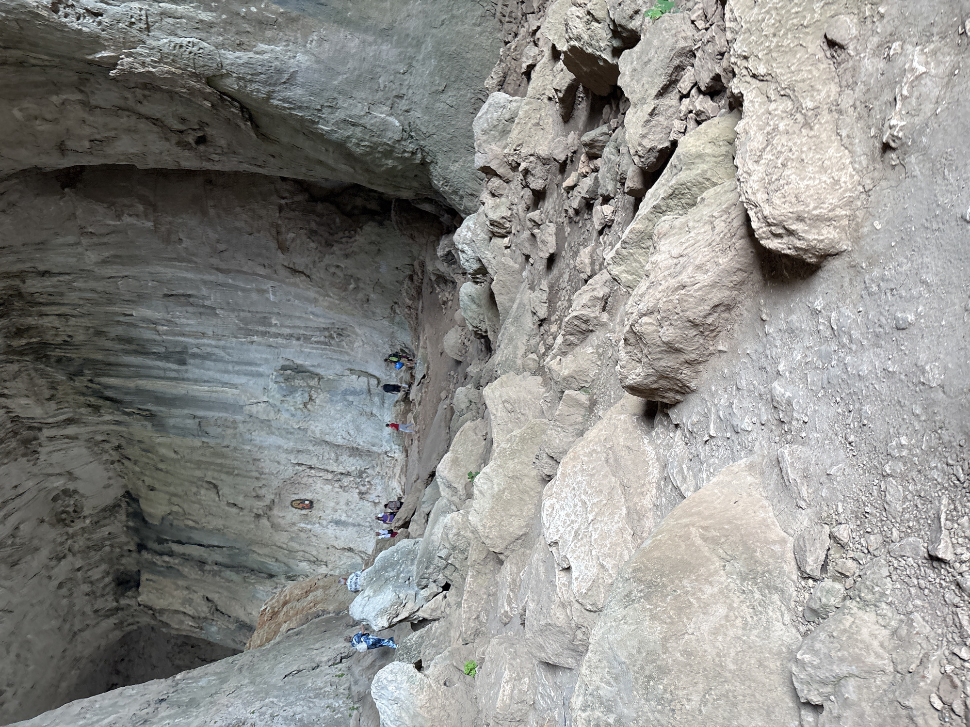 The image depicts a cave structure in Karlukovo, Bulgaria. The left side of the image shows the smooth, layered rock of the cave wall, with climbing holds affixed to it. Several climbers are visible on the wall, wearing various colors of clothing and climbing gear. The right side of the image shows a more rugged, rocky terrain. The lighting suggests the photo was taken during the daytime.