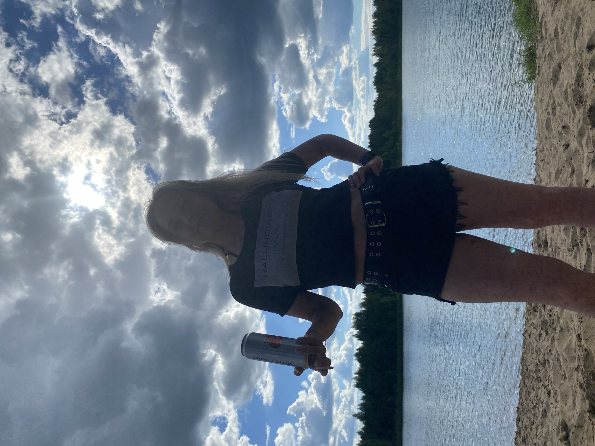 A woman stands on a sandy beach, holding a can of Stella Artois and a cigarette. She is wearing a black crop top with "BALENCIAGA HARLEM" printed on it and distressed black shorts. She is also wearing a black belt with double grommets and a dark watch. The woman is silhouetted against a dramatic sky filled with clouds, with the sun partially obscured. Behind her, a calm blue lake is visible, with a dark green forest line in the distance. The location is the city of Novyi, Russia.