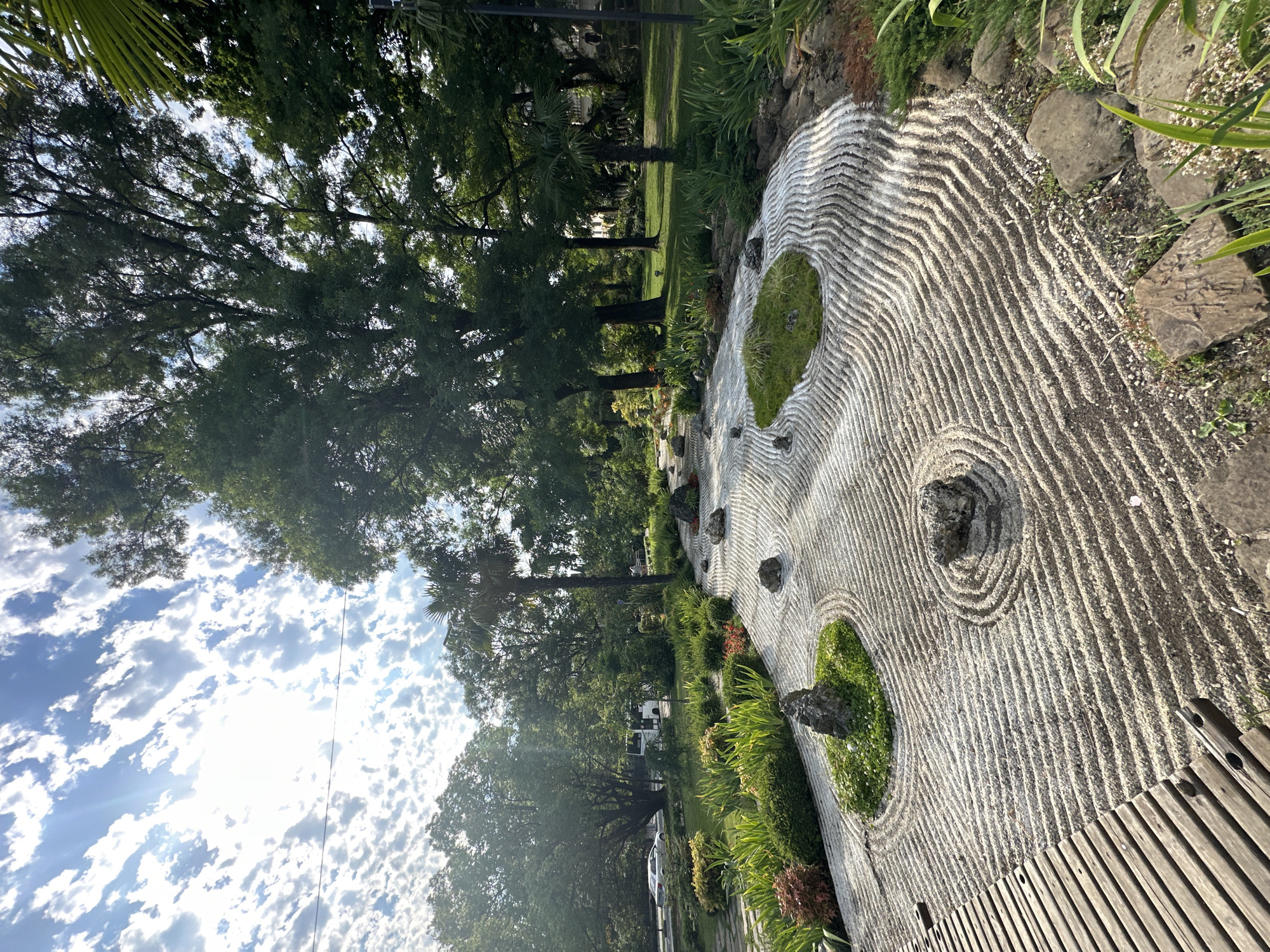 The image depicts a Zen garden within the Arboretum Botanical Garden in Sochi, Russia. The garden features a raked white gravel area with concentric wave patterns, interspersed with several dark rocks of varying sizes. Patches of green moss or grass are also present within the gravel. To the left, a wooden deck with horizontal planks is visible. Surrounding the Zen garden are lush green plants, shrubs, and trees. In the background, a road with a white car and a white bus can be seen, along with more trees and buildings. The sky above is blue with scattered white clouds, and sunlight is filtering through the leaves of the trees. The scene is captured in daylight, with shadows indicating a sunny environment. There are no people visible in the image.