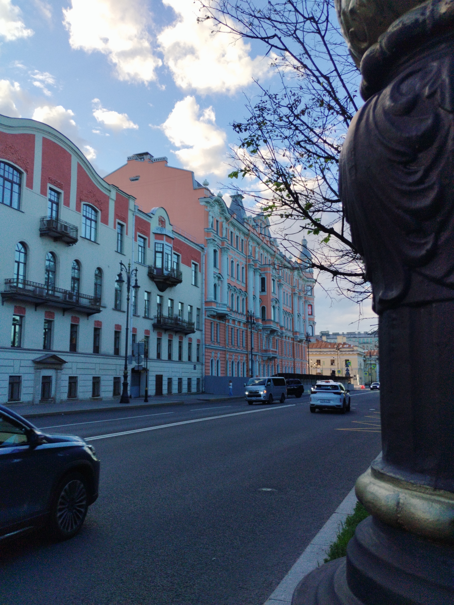 A street scene in Saint Petersburg, Russia, unfolds along a wide asphalt road lined with ornate, pastel-colored buildings. The buildings feature intricate architectural details, including arched windows, balconies with wrought-iron railings, and decorative cornices. A light pole stands in front of one of the buildings. Several cars are visible on the road, including a dark SUV in the foreground on the left, a silver van, and a white sedan in the middle ground. The sky above is a mix of blue with scattered white clouds. In the right foreground, a large, dark, textured sculpture, possibly part of a monument or lamp post, partially obstructs the view of the street and buildings. A bare tree branch extends into the frame from the upper right, silhouetted against the sky. The overall impression is of a European city street in the late afternoon or early evening.