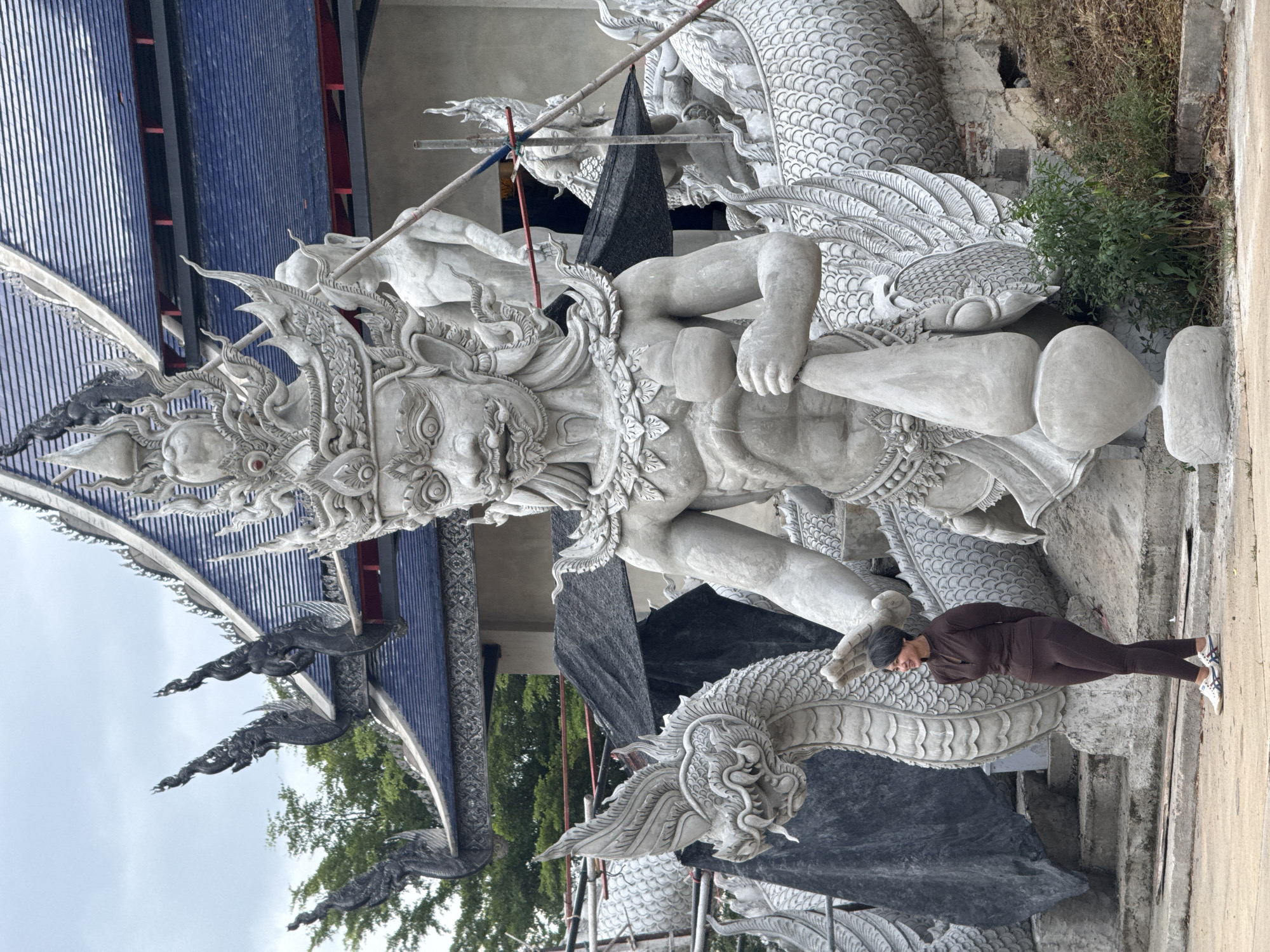The image depicts the exterior of a temple construction site in Cha-am, Thailand. The primary subjects are large, unfinished concrete sculptures of mythological figures, including a multi-headed serpent and a formidable humanoid entity with an elaborate headdress, positioned in front of a temple building with a blue tiled roof. A woman in a brown athletic outfit stands to the left of the sculptures, looking towards the right. Construction materials and scaffolding are visible around the sculptures and the building. The environment appears to be outdoors, with a cloudy sky and some greenery in the background.