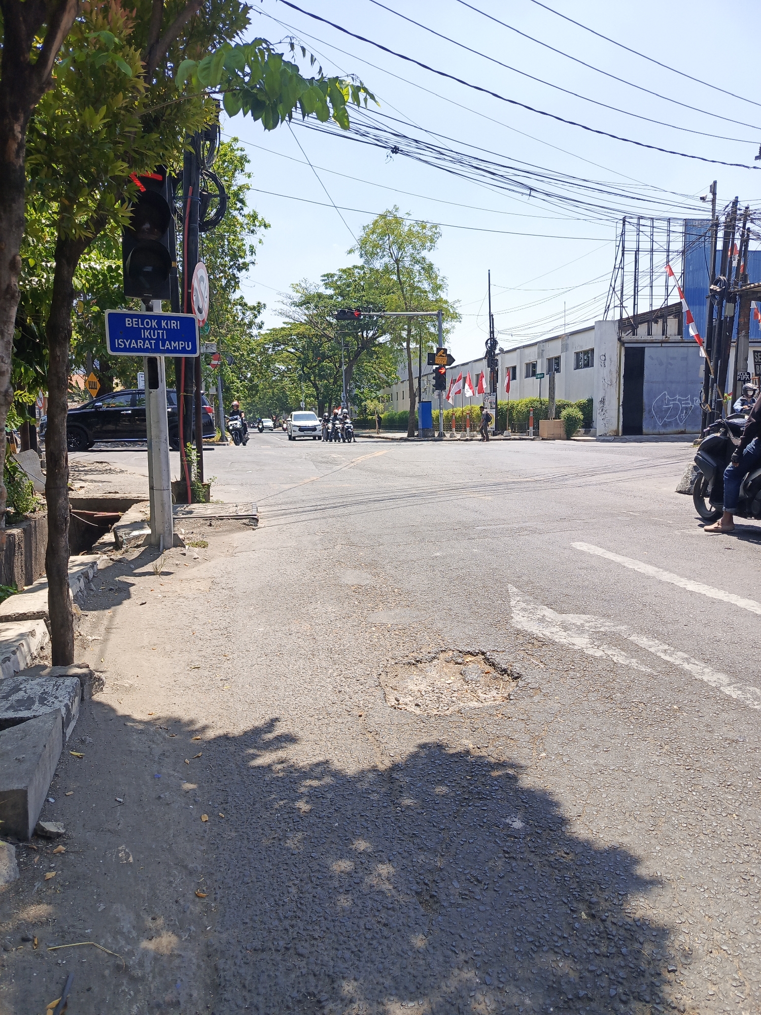 This image captures a street scene in Surabaya, Indonesia, likely near an intersection. The foreground is dominated by an asphalt road with a large pothole. To the left, a sidewalk runs along a tree-lined area, with a concrete curb. A blue street sign reads "BELOK KIRI IKUTI ISYARAT LAMPU," which translates to "Turn left follow the traffic signal." A traffic light pole with a red light illuminated stands nearby.
In the background, the intersection is busy with several motorcycles and a white car. Beyond the intersection, there's a building with some Indonesian flags displayed. Power lines crisscross overhead. The sky is clear and blue, indicating a sunny day. The overall environment suggests a typical urban setting in Indonesia.