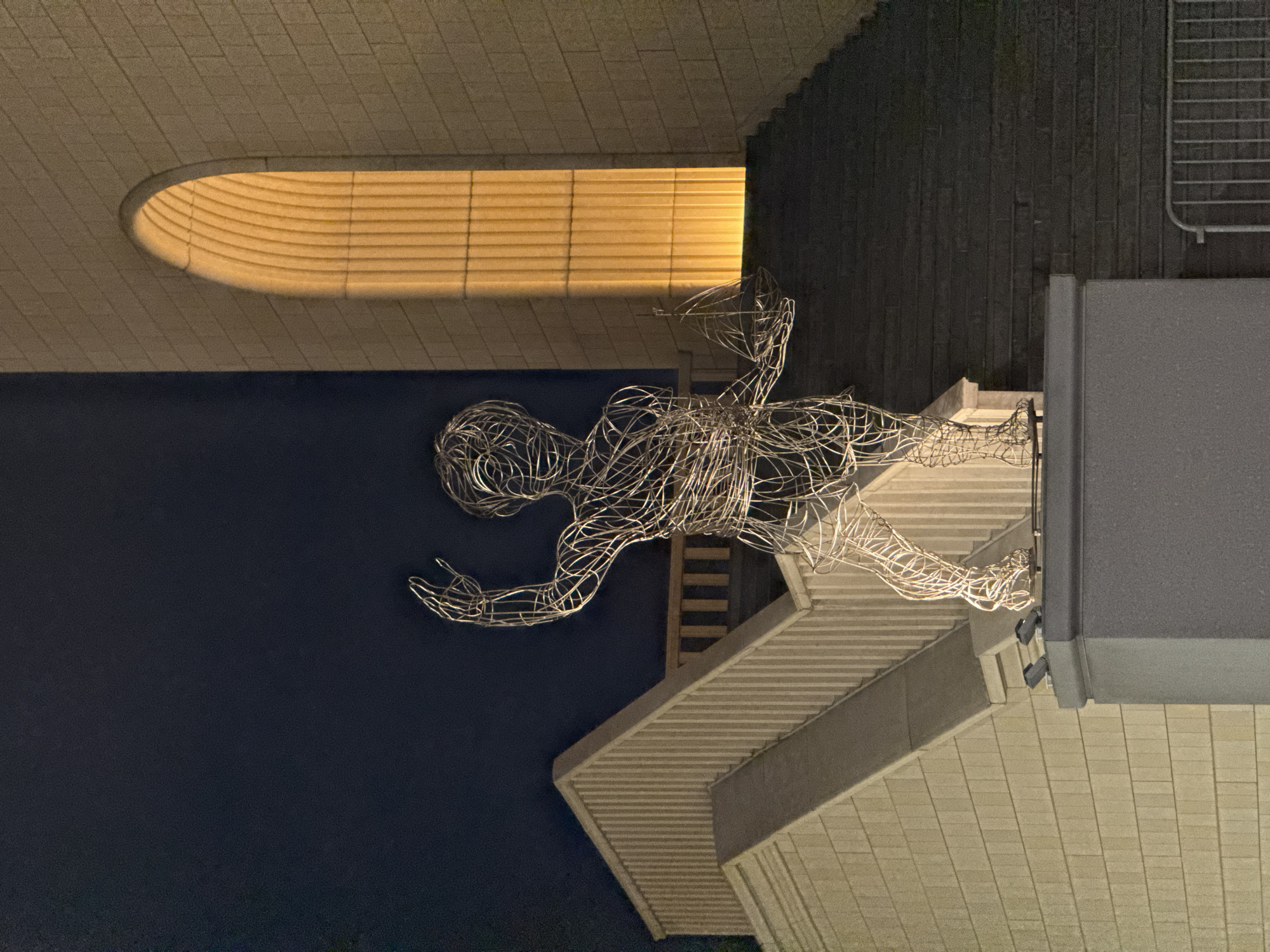 The image shows a sculpture of a human figure made from wire, situated on or near the roof of a building. The figure is positioned as if in motion. The background features architectural elements including stairs, a wall with vertical stripes, and a dark, solid-colored area. There is also a section with horizontal lines and an oval opening with a light source behind it. The image appears to be taken at night, as it has limited lighting. The location is Moscow, Russia.