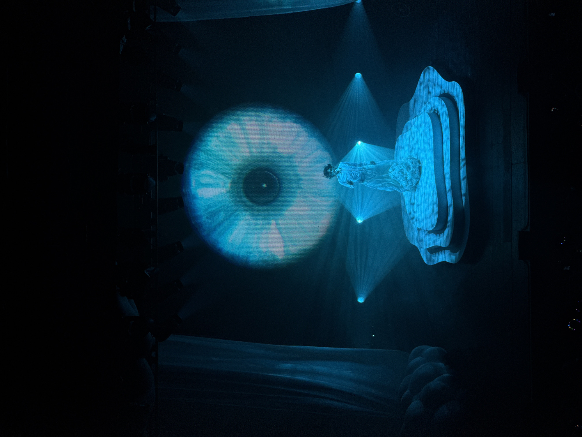 A stage scene, likely a performance art piece, bathed in blue light. A large projection of an eye is on the left side. Emanating from the right are spotlights that illuminate a figure lying horizontally inside a multi-layered, ornate frame. The lower part of the stage has a curved, abstract form that could be a prop or part of the stage design. The location may be in the city of Los Angeles, United States of America.