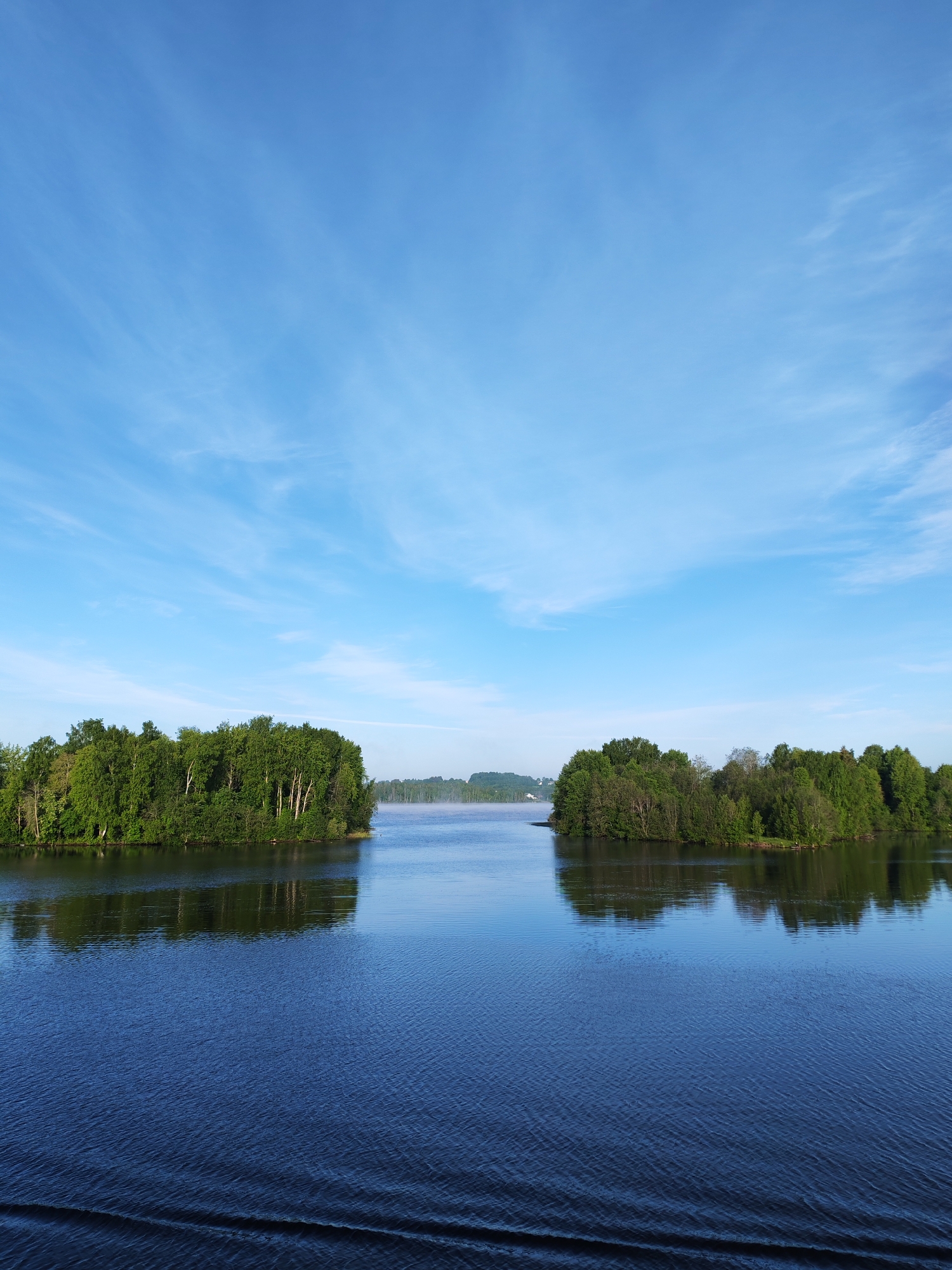 The image depicts a body of water, possibly a river or lake, with two densely forested islands on either side. The water is dark blue, and ripples are visible on its surface. The sky is a light blue color with wispy clouds. The overall environment is serene and natural. The location is likely near Irma, Russia, given the request's inclusion of the city's name, but this cannot be definitively confirmed from the image alone. There are no people visible in the scene. The primary subjects are the water, islands, trees, and sky. The only action implied is the natural movement of the water and clouds.