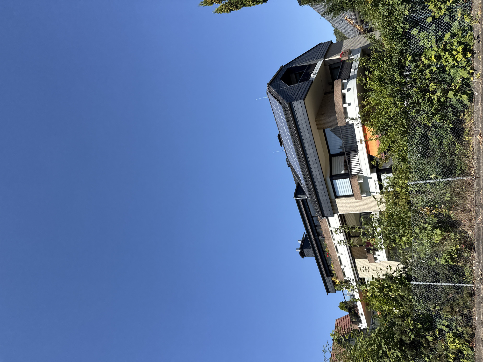 The image depicts a modern apartment building situated in Bad Sachsa, Germany, under a clear, vibrant blue sky. The building, constructed with light-colored brick and dark gray roofing materials, features multiple balconies and a sloped roof equipped with solar panels. Lush green foliage and trees surround the property, with a chain-link fence in the foreground separating the building from the viewer's perspective. The scene is quiet and static, with no people or ongoing activities visible. The lighting suggests a sunny daytime setting, likely late morning or afternoon.