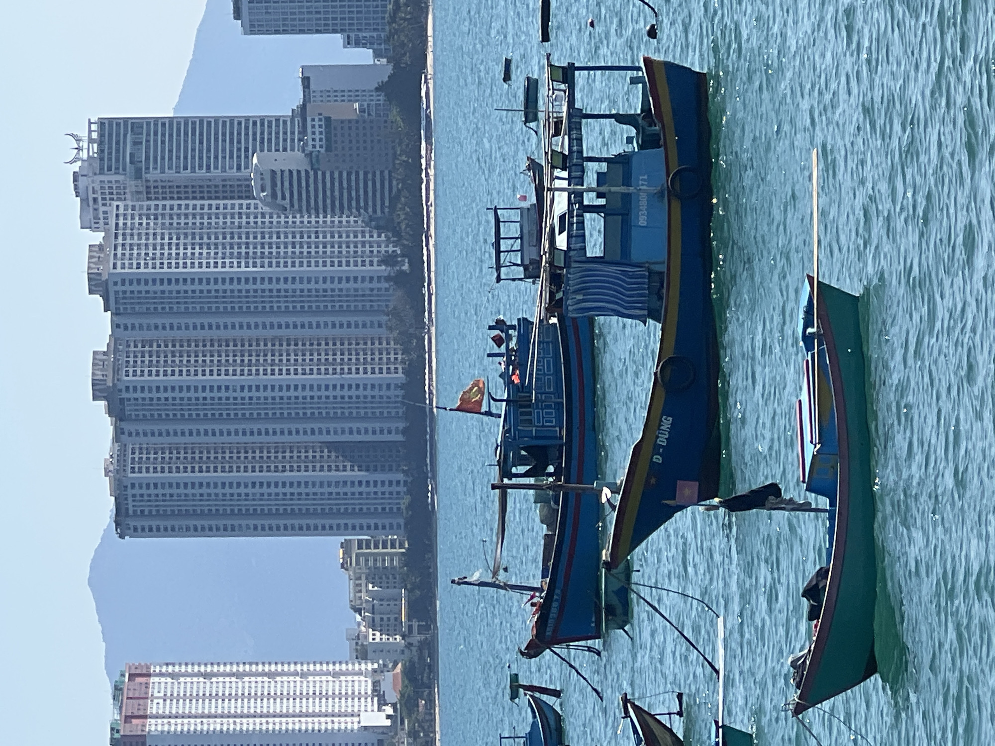 The image displays a maritime scene in Nha Trang, Vietnam, under a clear, light blue sky. The foreground and midground feature multiple traditional Vietnamese fishing boats moored on calm, turquoise-blue water. These vessels primarily exhibit blue hulls with yellow, red, or green trim. Identifying markings include "D-DUNG" on one boat's bow and numbers such as "093480171" and "KA1038678" on others. One larger boat displays a Vietnamese flag from its stern, and several possess rudimentary canopies or cabins. No individuals are visible on or immediately adjacent to the boats, which are stationary.
The background reveals a dense urban coastline. Numerous modern high-rise buildings, predominantly multi-story residential or hotel structures, dominate the skyline. One building on the left exhibits red construction scaffolding or framing at its uppermost levels. A prominent high-rise in the center features a distinct, wing-like architectural element on its roof. A narrow strip of sandy beach is visible along the coast, separating the water from the urban structures. Distant, hazy blue mountains delineate the furthest background.