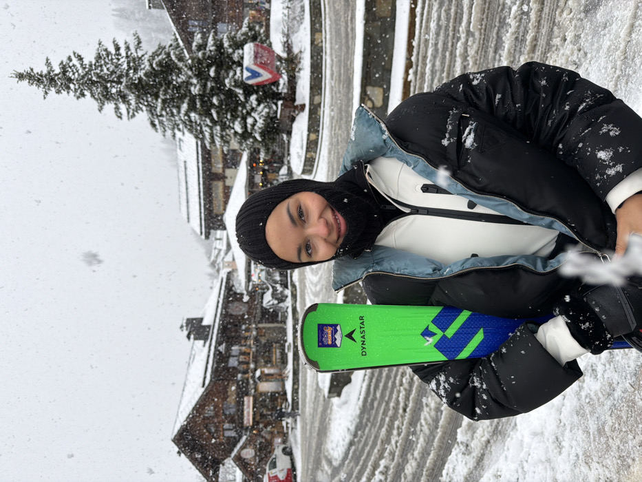 A young woman is depicted in a close-up, waist-up portrait during a snowfall in Les Allues, France. She faces forward, smiling, wearing a black ribbed balaclava, a light-colored zippered jacket, and a black puffer jacket with a light blue collar. Snowflakes are visible on her clothing. She holds a vertical, bright neon green Dynastar ski with blue and white graphics on its side, positioned to the left of her body. Her hands are gloved, though partially obscured.
The background shows a snow-covered village street or square. To the left, several multi-story, dark-wood chalet-style buildings with snow-covered roofs are present, some with illuminated windows. A red vehicle is partially visible on the far left. To the right, a large, snow-laden coniferous tree stands prominently. At its base, there is a low stone wall or planter with a distinct blue, white, and red sign or emblem. The ground is covered in a mix of compacted snow and slush, showing patterns consistent with vehicle or foot traffic. The sky is overcast and bright, contributing to the even light.