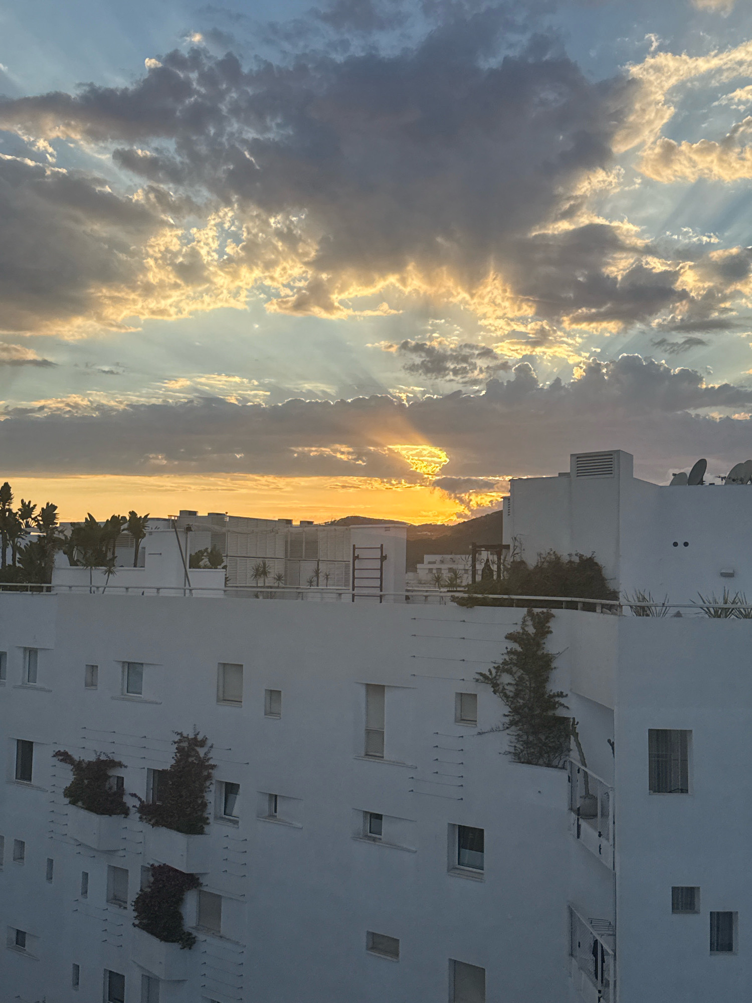 The image captures a sunset over a modern, whitewashed building complex in Ibiza, Spain. The sky is filled with dramatic clouds, with rays of sunlight breaking through, casting a warm glow on the scene. Palm trees are visible in the background, adding a tropical touch. The building features balconies with greenery, and satellite dishes adorn the rooftop, typical of Mediterranean architecture. The overall atmosphere is serene and picturesque, highlighting the beauty of an Ibiza sunset.