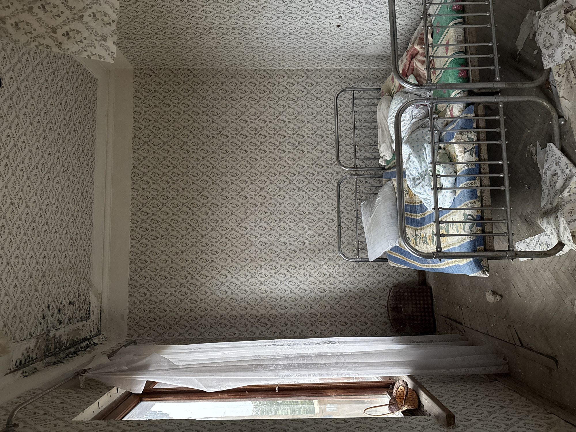 An interior view in Tskaltubo, Georgia, reveals a deteriorated bedroom. Two metal-framed beds stand adjacent to each other, covered with bedding and other items. The wallpaper is patterned and exhibits water damage and mold growth along one wall. A window is draped with a white curtain, and a basket rests on the windowsill. The floor is wooden and visibly worn. A small, woven basket is positioned near one of the beds.