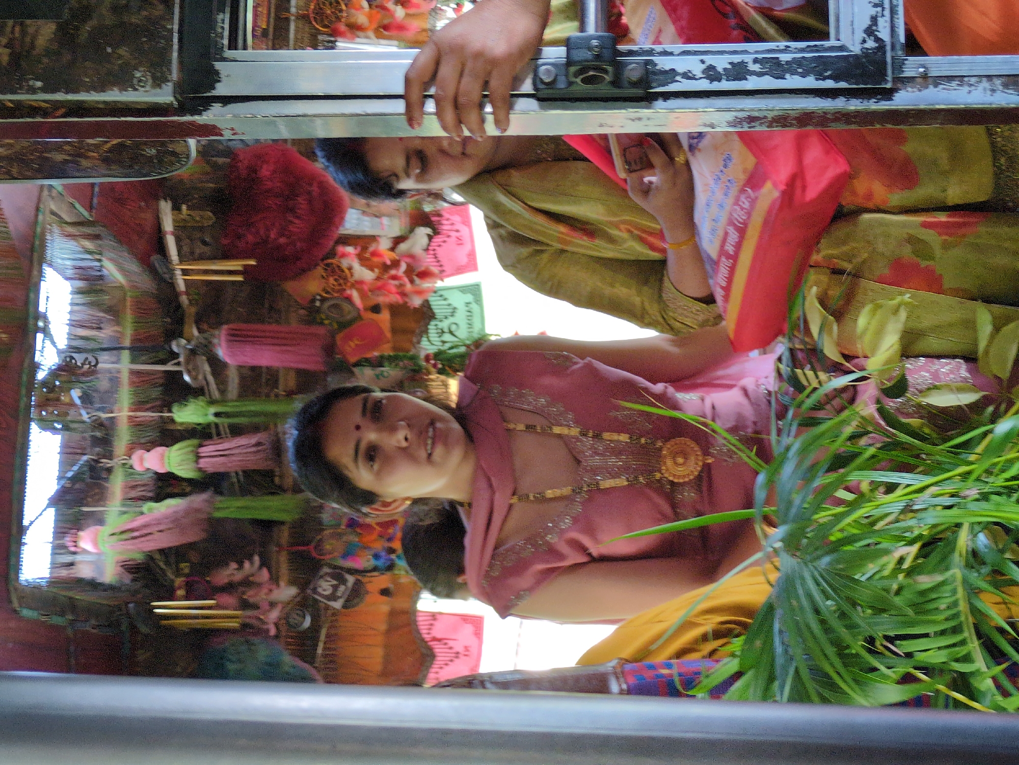 The image captures a scene inside what appears to be a mode of transport decorated with various colorful tassels and ornaments. In the foreground, a woman wearing a pink outfit and an elaborate gold necklace is looking forward. To her right, another woman in a green and red patterned garment is visible, holding a red bag. The environment is cluttered with decorative items hanging from the ceiling, including tassels and possibly wind chimes. A large plant with green leaves is positioned in the lower left of the frame. The scene suggests a festive or devotional setting, possibly a decorated bus or van, within the city of Gumma, India.