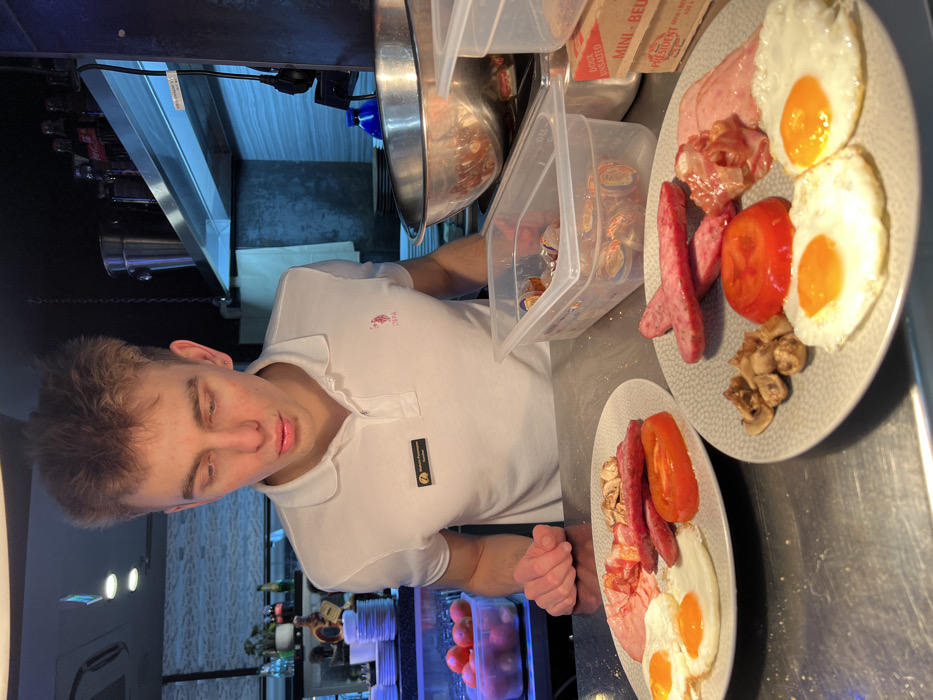 A young adult male, identified by a name tag as "Mihailo Jovanović, Kuvar" (Chef), stands behind a stainless steel counter in what appears to be a commercial kitchen or serving area. He has short, light brown hair and wears a white U.S. Polo Assn. polo shirt. His gaze is directed downward towards two identical plates of breakfast food on the counter. Each plate contains two sunny-side-up fried eggs, slices of cooked sausage, pieces of cooked bacon, slices of ham, cooked tomato slices, and sautéed mushrooms. His hands rest on the counter near the plates. In the background to the left, a clear container holds red fruit or vegetables, and stacks of white plates are visible. To the right, shelves hold a silver bucket, glass bottles including Coca-Cola, and a large metal mixing bowl. Clear plastic containers on the counter hold food items, one displaying foil-wrapped portions, and a carton labeled "President Mini-Beurre Doux Unsalted" is also present. The background wall features light blue or grey tiling under a bright overhead light. The specific location within the city of Budva, Montenegro, is not discernible from the image.