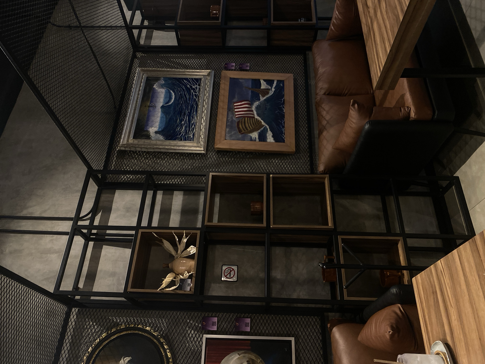 The image shows a dimly lit interior space, possibly a lounge or café, featuring a black metal frame structure that houses decorative elements and furniture. The structure includes shelves with framed artwork, such as ocean wave paintings and a depiction of a sailboat with an American flag. On one shelf, there's a decorative object that resembles a corn cob with leaves. The shelves also hold small decorative boxes and a "no smoking" sign.
Adjacent to the shelving unit, a brown leather couch or seating area is positioned with a dark-colored cushion. A wooden table is situated next to the seating area. The wall behind the shelving unit is a textured gray, possibly concrete. The overall arrangement gives the impression of an eclectic and stylized design.
