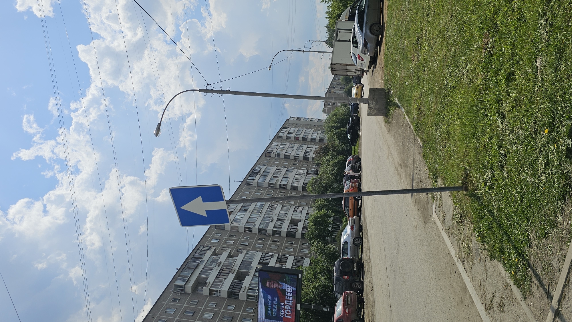 The image shows a street scene in an urban environment. A tall residential building with multiple windows and balconies is visible in the background. Several parked cars line the side of the street. A blue directional sign with a white arrow pointing to the left is prominently displayed on a pole. A billboard advertising "СЕРГЕЙ ГОРДЕЕВ" (Sergei Gordeev) is also visible. Overhead power lines stretch across the sky filled with clouds. Grass grows along the edge of the street.
