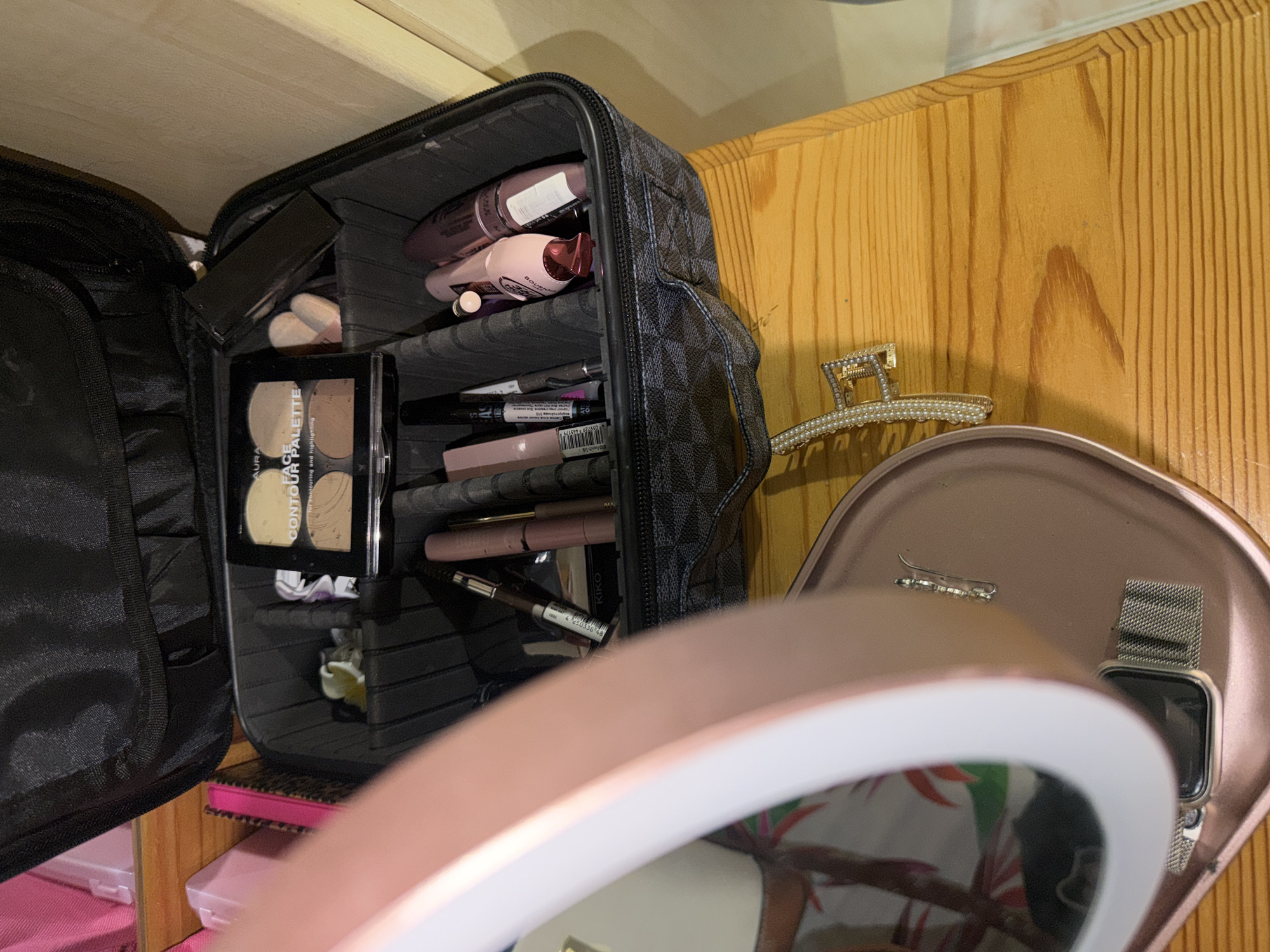 The image displays an organized collection of cosmetics and personal accessories, likely situated on a wooden vanity or shelf indoors. In the center, a black and gray patterned makeup bag, open, reveals several compartments filled with beauty products. Visible items inside include pink and dark-colored mascara tubes, one of which is from Bourjois. An "AURA CONTOUR FACE PALETTE" for "contouring and highlighting" is prominent, along with other makeup brushes and pens, including a KIKO product with a visible barcode (002624 40071).
To the right, a gold-toned hair claw clip adorned with pearls rests on the wooden surface. Partially in the foreground, a rose gold-colored mirror with an illuminated ring light is visible, reflecting an abstract pattern. Adjacent to the mirror, a rose gold-colored tray holds a silver ear cuff and a smartwatch with a silver mesh band. Below the makeup bag, portions of pink storage boxes or drawers can be seen. The scene is well-lit, typical of an indoor setting, with no specific activity taking place.
