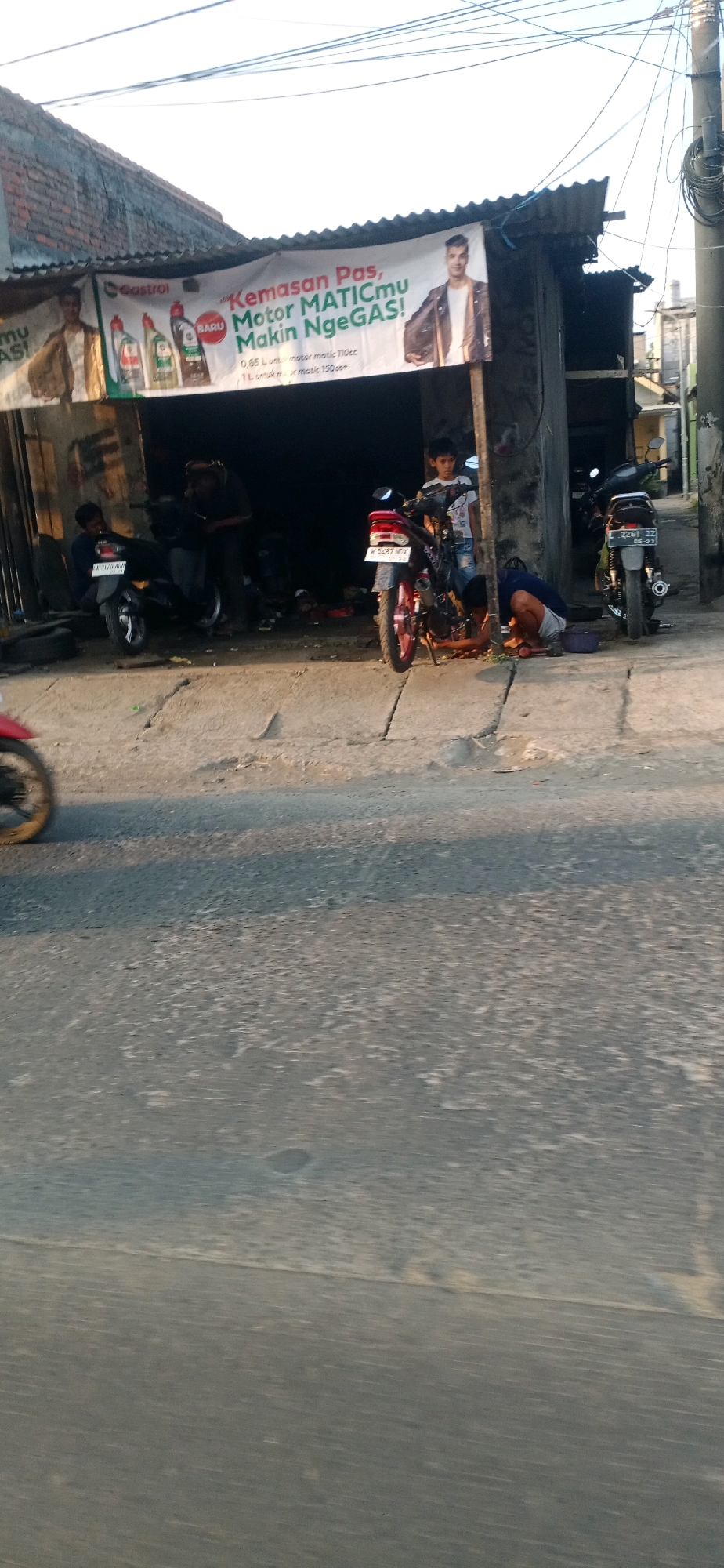 The image captures a street-level view of a small, open-air motorcycle repair shop in Surabaya, Indonesia. A prominent banner overhead advertises "Castrol" motorcycle oil with the slogan "Kemasan Pas, Motor MATICmu Makin NgeGAS!". The banner features images of oil bottles and two men.
In the foreground, a person on a red motorcycle is partially visible, suggesting movement or recent passage. The repair shop itself appears to be constructed from corrugated metal and wood. Several motorcycles are present, including one being worked on by a person kneeling beside it. Another person, possibly a young boy, stands near this motorcycle. Further inside the dimly lit shop, more motorcycles and general workshop clutter are visible. To the right, a second motorcycle is parked, and another person is seen crouching. The street in front of the shop is paved and appears to be somewhat weathered. The overall impression is of a functional, everyday scene in a bustling urban environment.