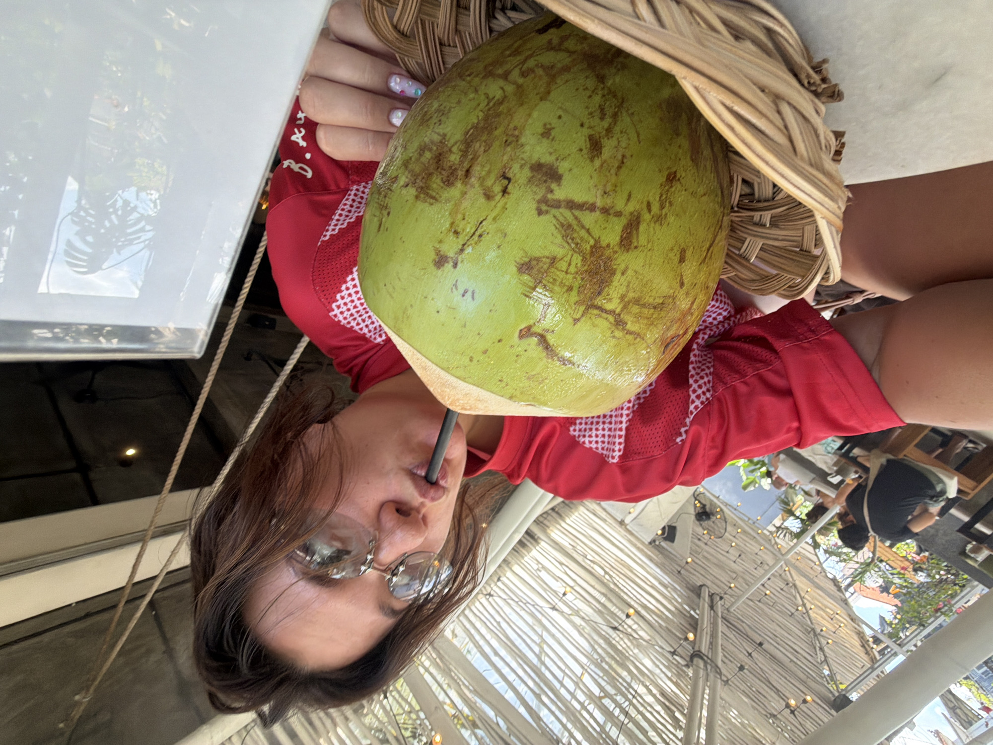 In North Kuta, Indonesia, a young woman with dark hair wears round sunglasses and a red athletic shirt. She is leaning forward, sipping from a large green coconut through a black straw. The coconut rests in a woven basket. The background shows a covered outdoor seating area with a slatted roof, strings of lights, and visible foliage, suggesting a restaurant or cafe setting. The woman's right hand, adorned with colorful nail art, is near the coconut.