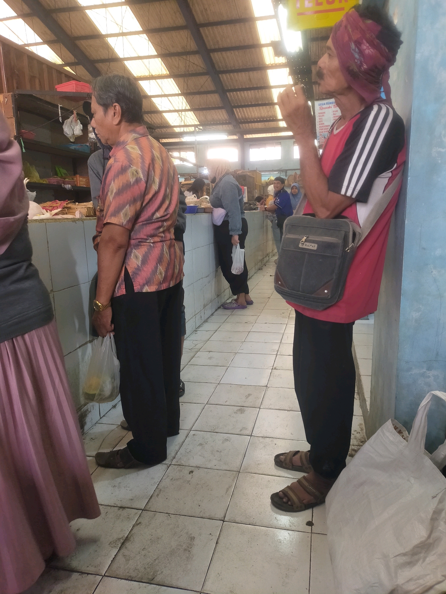 The image depicts a street market scene in Bululawang, Indonesia. In the foreground, a man stands near a blue wall, wearing a red t-shirt with black and white stripes on the sleeves, black pants, and brown sandals. He has a bandana wrapped around his head and is holding a cigarette. A dark-colored messenger bag is slung across his chest. To the left, a person in a pink pleated skirt and a dark grey top is partially visible. Further in the background, several other individuals are present, some appearing to be vendors or shoppers. The environment suggests an open-air market with a tiled floor and a corrugated metal roof supported by wooden beams. Merchandise and bags are visible on the counter and on the floor.