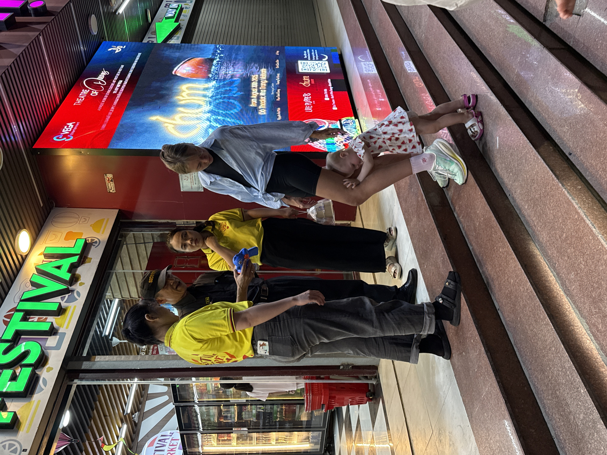 The image shows a group of people in what appears to be an urban setting, possibly in Vietnam given the location context. In the foreground, two people wearing yellow t-shirts are visible, and another person in a security uniform is talking to them. They are standing in front of a store with a large, brightly lit sign that says "FESTIVAL" in green, stylized letters. Behind them, a large LED screen displays advertisements for a show called "Chum" from "THEATRE OF DREAMS," produced by VEGA Entertainment. The screen also shows text in what appears to be Vietnamese and Russian, indicating show dates and location: "From August 30th 2025 Do Theater, Nha Trang Vietnam."
Further up the stairs, a woman is holding a baby. She is wearing a light blue shirt and dark shorts, while the baby is dressed in a patterned onesie. The woman's legs are extended, and she is holding the baby over her lap. Other individuals' legs and feet are visible on the stairs, some wearing sandals and sneakers. The stairs themselves are made of reddish-pink speckled stone. The overall scene suggests a public space, possibly an entertainment district or a plaza, with the activity of people interacting and the backdrop of advertisements. The lighting indicates it is likely evening or nighttime.