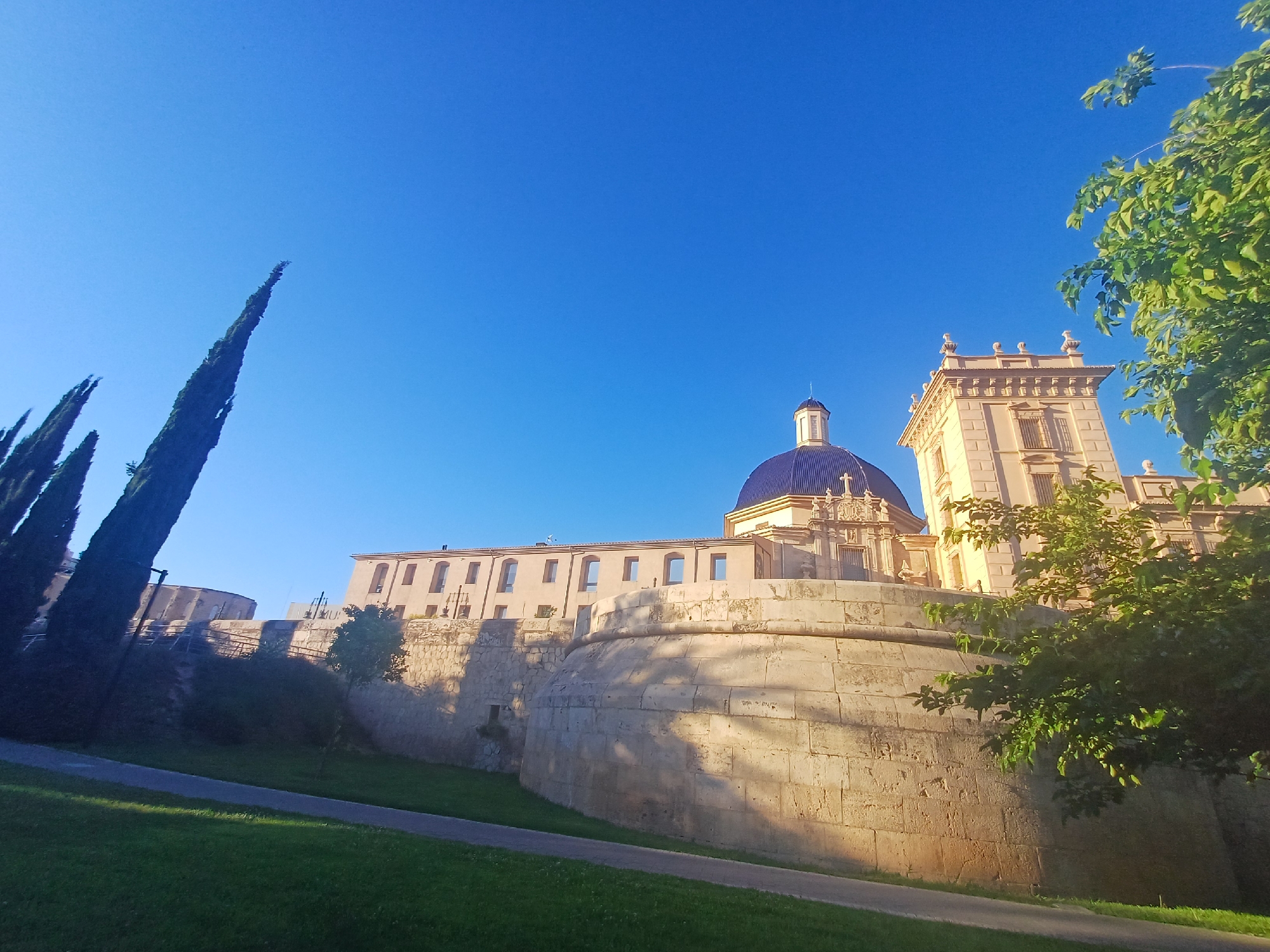 The image shows a view of the Museo de Belles Arts de València, located in València, Spain. The museum's facade features a rounded stone wall and a section of the building with arched windows. A blue-domed structure is visible behind the main building. Adjacent to the museum is a square tower-like structure with decorative details. The foreground shows a grassy slope and a paved pathway. Tall, slender trees rise from the left side of the frame, and lush green foliage fills the right side. The sky is clear and blue.