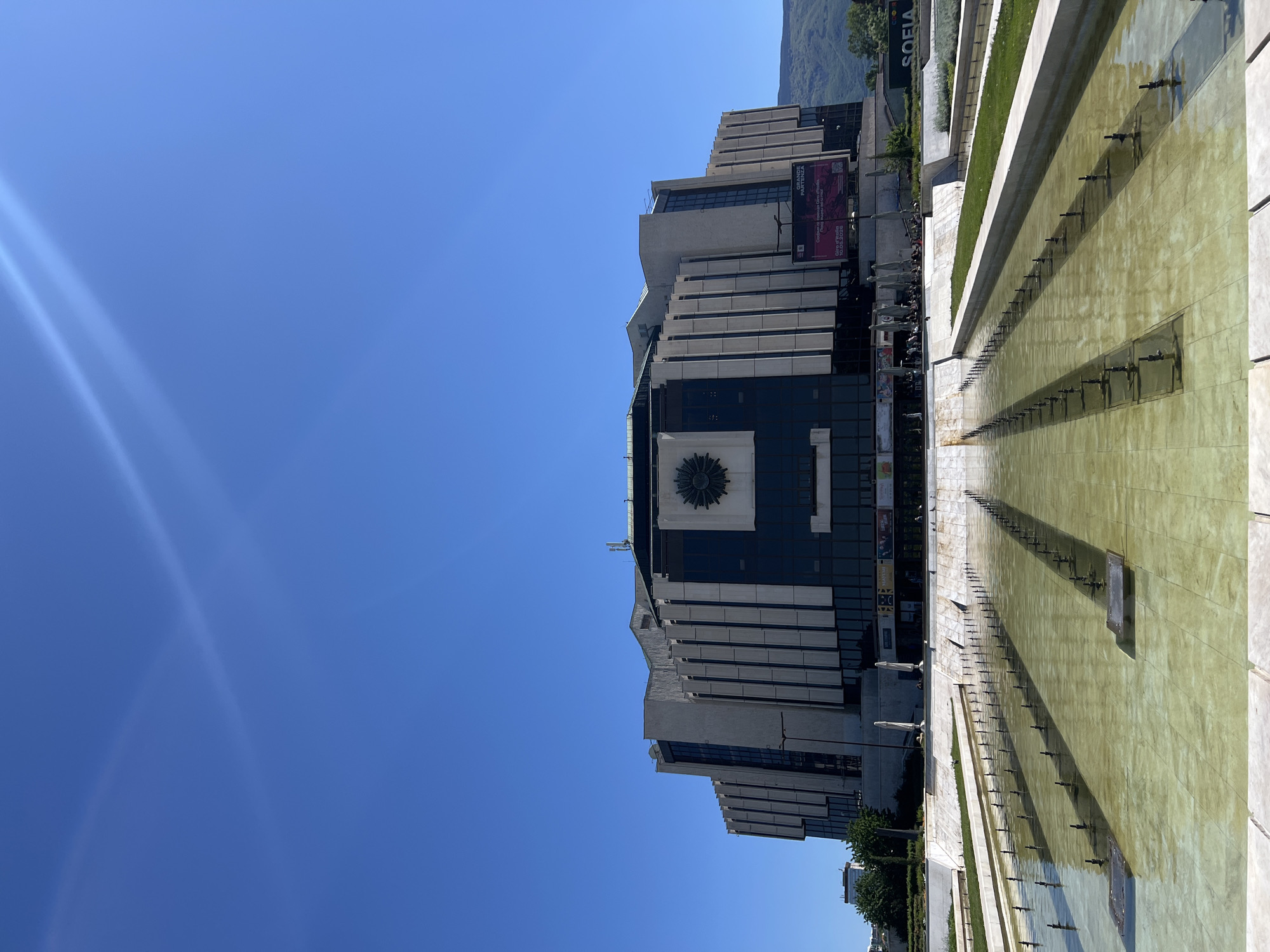 The image shows the National Palace of Culture in Sofia, Bulgaria, viewed from across a long, shallow reflecting pool with numerous fountain nozzles. The building has a multi-tiered, modernist design with a prominent central emblem. Several people are present in front of the building, along with banners and posters. The sky is clear and blue. The pool surface reflects light and the building's silhouette. A mountain is visible in the background. The word "SOFIA" is visible on a sign near some vegetation in the background.