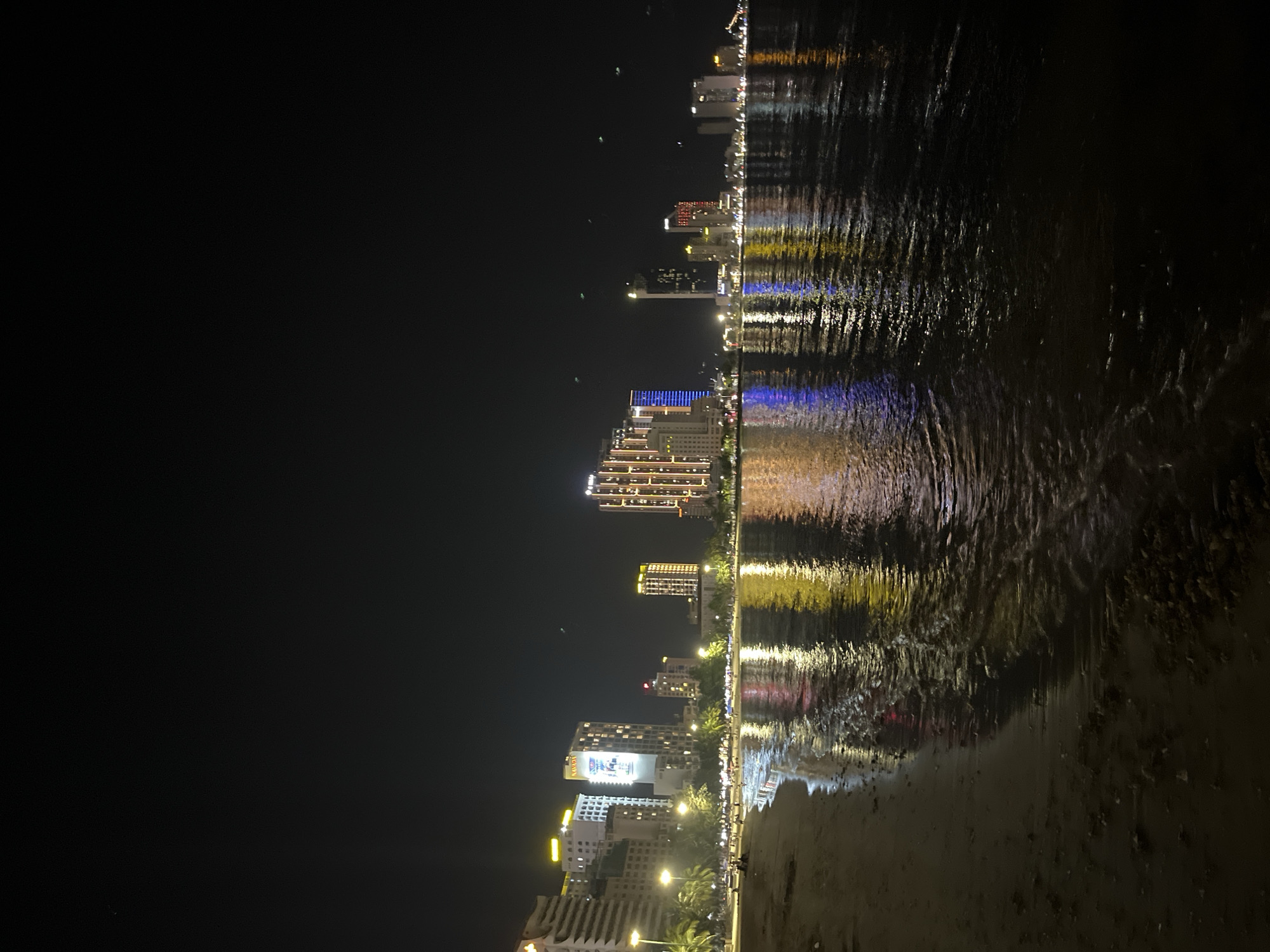 The image captures a nighttime view of a city skyline reflecting in water, likely a sea or bay. The buildings are illuminated with various colors, creating a vibrant reflection in the water. The environment appears to be a shoreline or beach, given the presence of sand or dark material at the bottom of the frame. The overall scene suggests an urban coastal area at night, with the city lights serving as a focal point.
