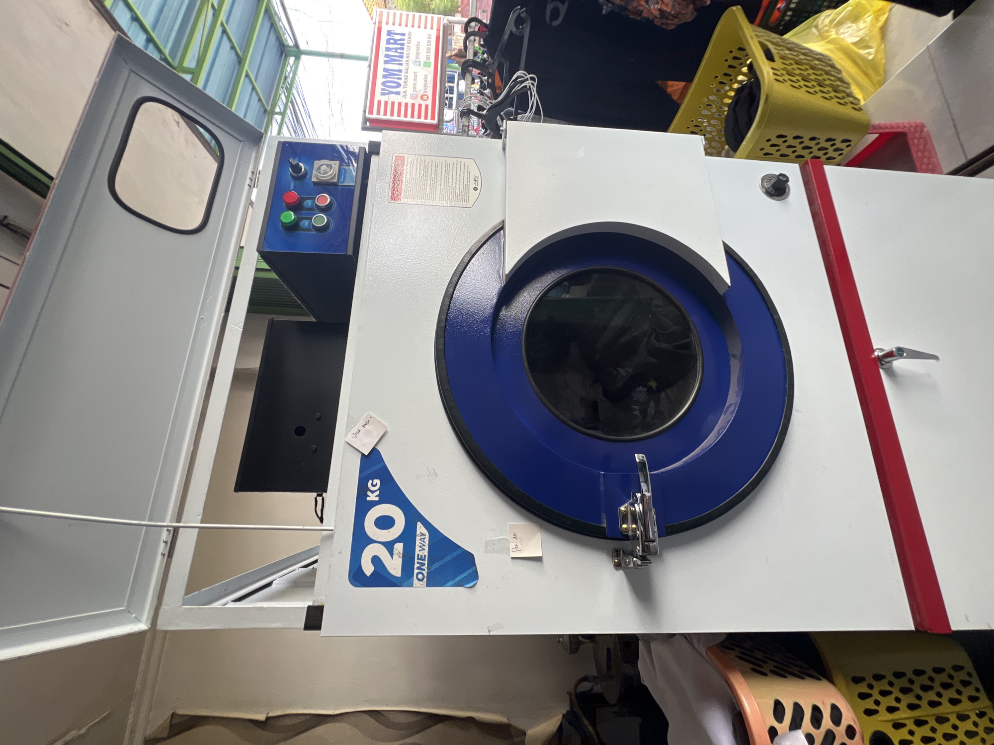 A commercial washing machine, possibly a dryer, with a 20 KG capacity and "ONE WAY" branding is visible. The machine is white with a prominent blue circular rim around the drum opening, which is closed. A silver handle with a latch mechanism secures the door. Above the drum, a blue control panel with red and green buttons and a switch is present. To the right of the machine, a sign for "YOM MART" is displayed, indicating its location on Jln. Tukad Balian No. 135, Renon, Denpasar, Indonesia. The sign also features social media icons and a phone number. In the foreground, laundry baskets, one yellow and one orange with a white cloth, are placed to the left of the machine. Several black clothes hang on hangers to the right, suggesting the area is a laundromat or dry cleaner. The background reveals a covered outdoor area with a corrugated plastic roof.