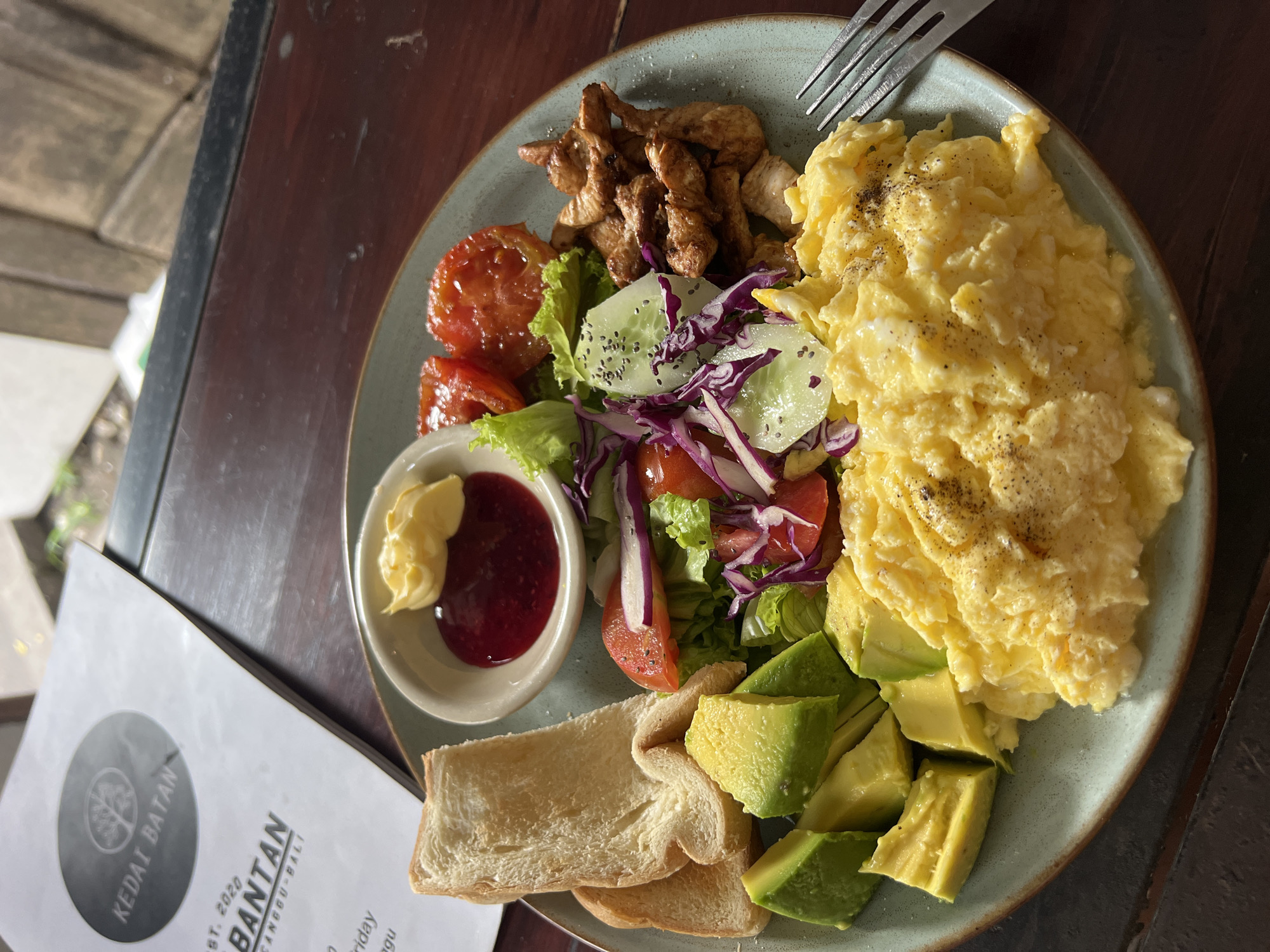 A plate of breakfast food is seen on a wooden table, with a piece of paper next to it identifying the location as Kedai Batan, Canggu-Bali, North Kuta, Indonesia. The plate contains scrambled eggs, chicken pieces, a side salad, avocado, toast, and a small bowl with jam and a dollop of yellow spread, possibly butter or mayonnaise. A fork rests atop the eggs. The meal appears to be eaten outdoors.