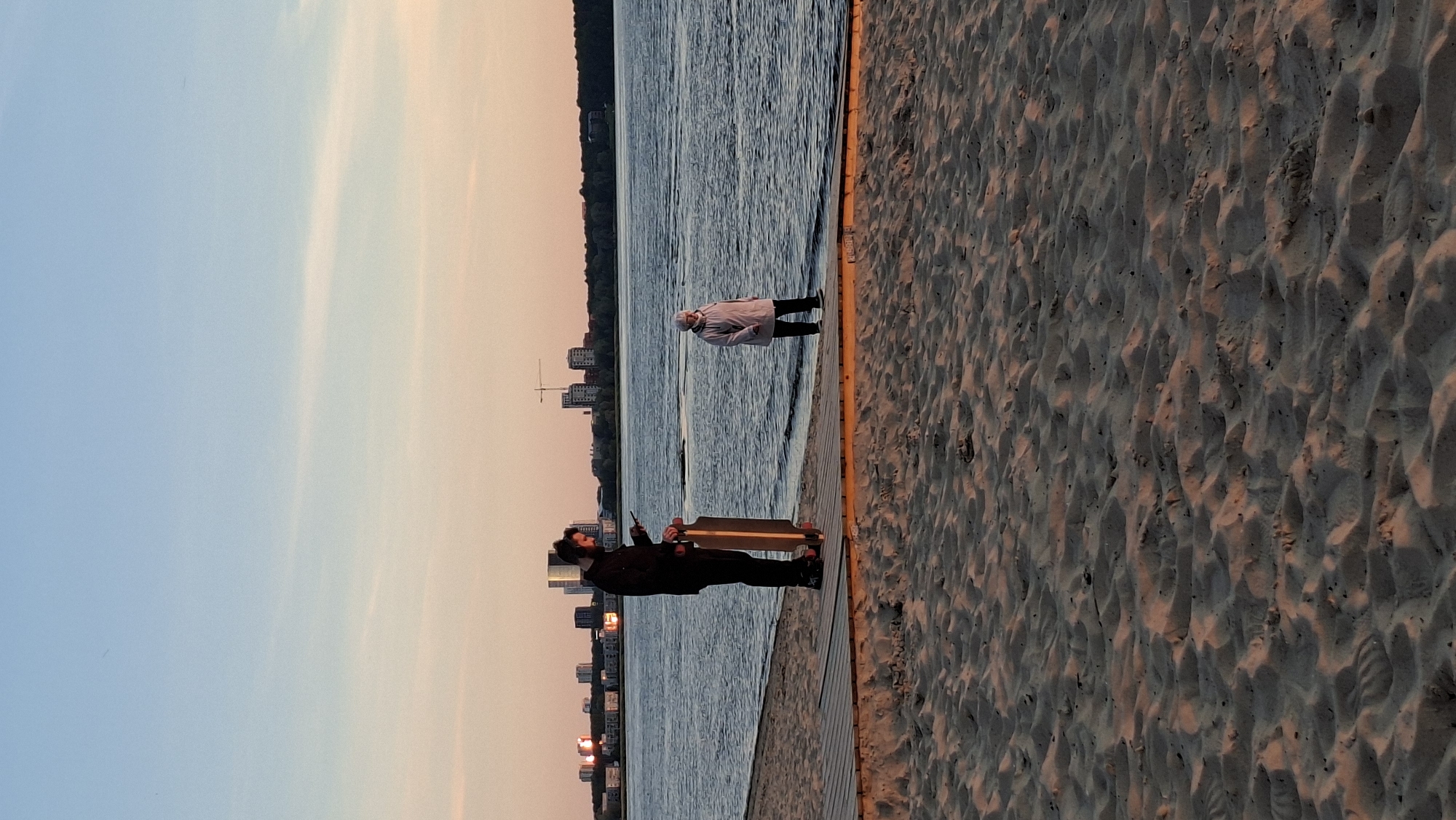 The image shows a sandy beach with a wooden boardwalk by the water. Two people are on the boardwalk. On the left, a man is standing and looking at his phone while holding a longboard. He is wearing dark clothing and headphones. On the right, a woman in a light-colored jacket and dark pants is standing and looking towards the water. In the background, across the water, there is a city skyline with buildings and a construction crane. The sky is clear with a gradient of light blue and soft orange, indicating it is likely sunset or sunrise. The scene appears to be in Tallinn, Estonia, with the city skyline characteristic of the area.