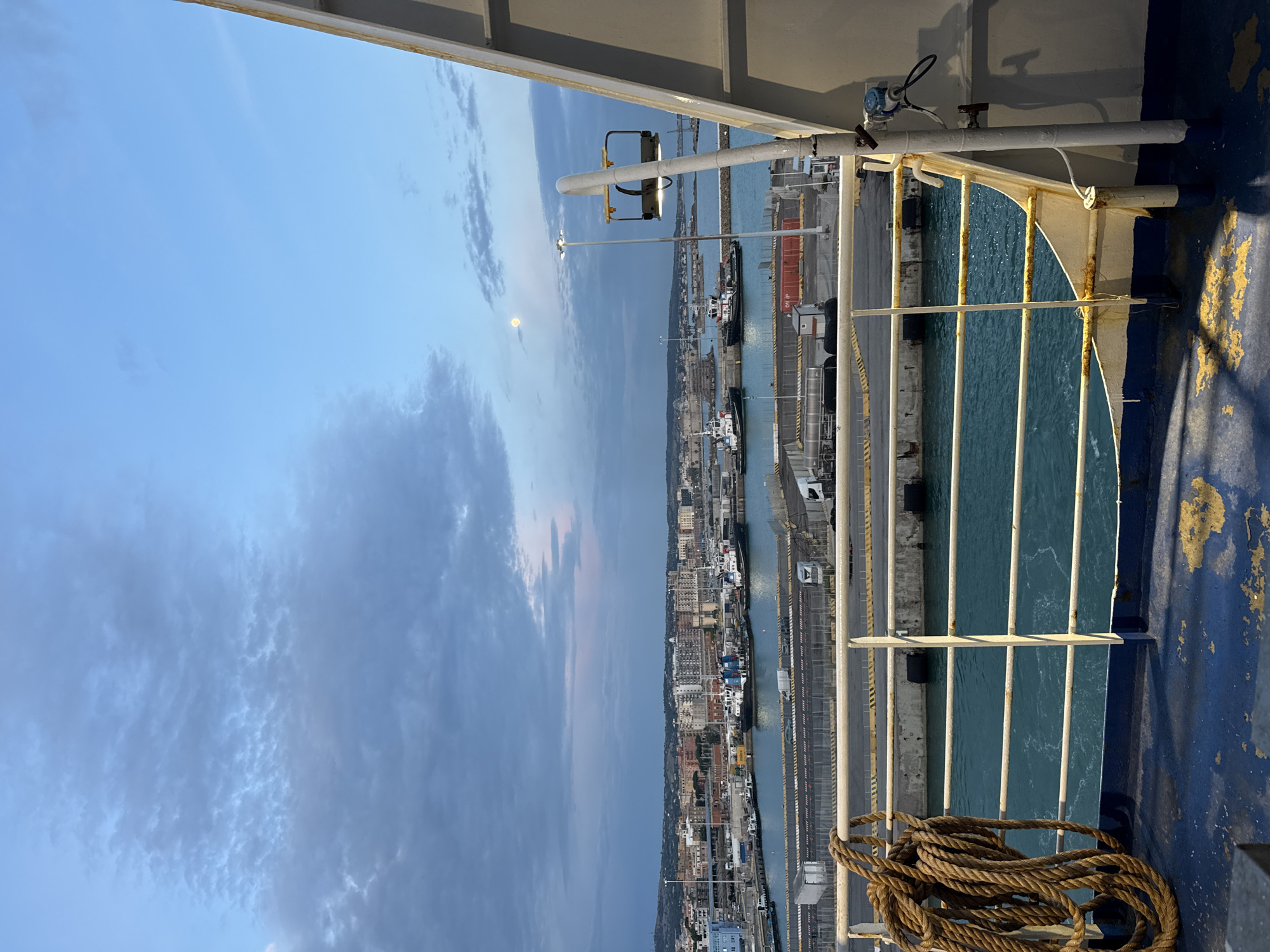 The image depicts a view from what appears to be a ship or ferry, overlooking the city of Civitavecchia, Italy. The sky is a mix of light blue and cloudy grey, suggesting either dawn or dusk. The city stretches along the coast, with buildings of varying heights and colors. A harbor is visible with several boats docked alongside concrete quays. Barriers line the edge of the quay. The sea is turquoise. The ship's railing is on the right side of the image, with a coiled rope in the foreground. Metal and painted structures are visible.