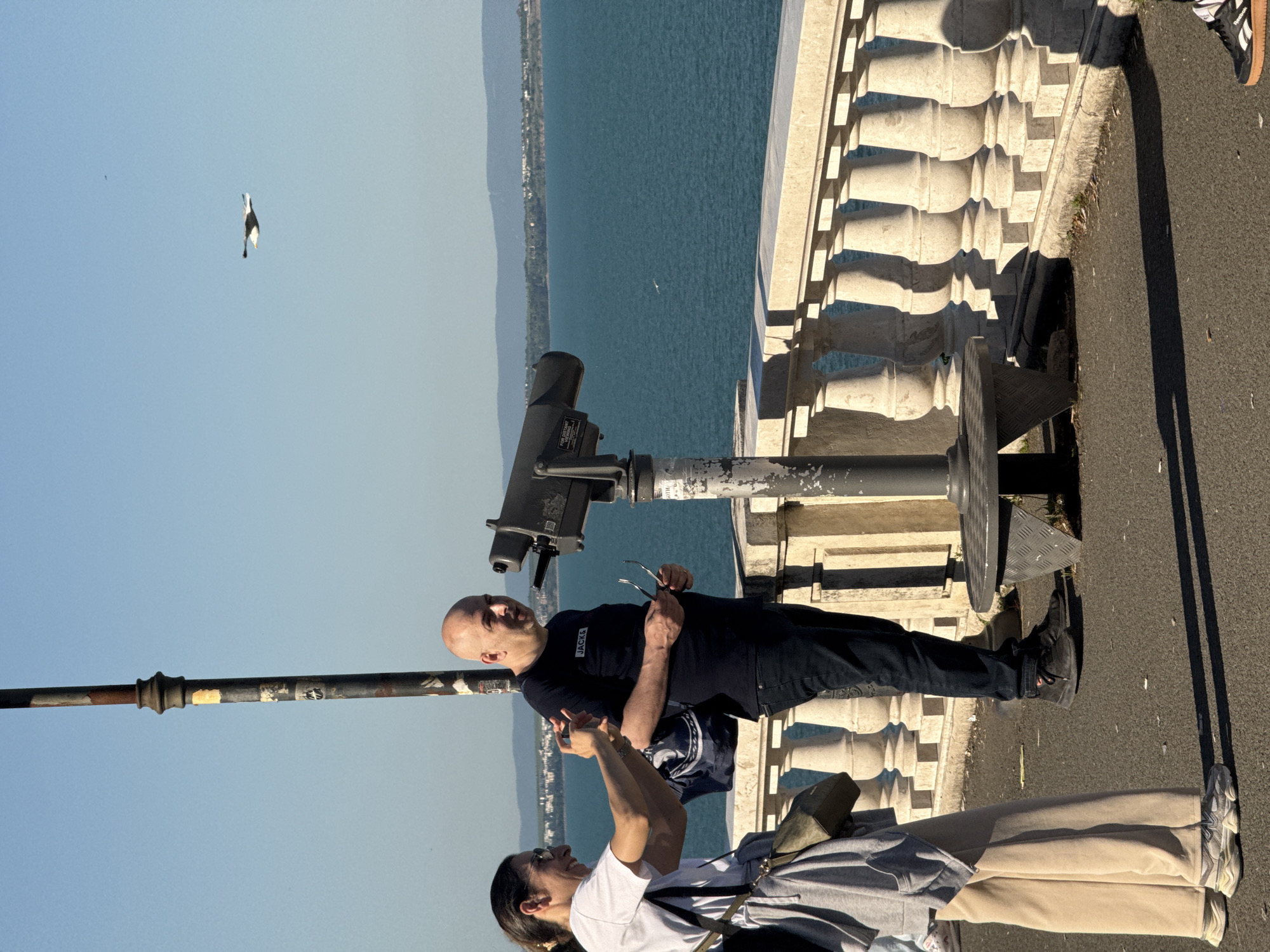The image shows two people at a seaside observation point. A woman with sunglasses is taking a photo with her smartphone, and a bald man in a black shirt holds his own glasses in his left hand. They are standing next to a coin-operated telescope fixed on a metal stand and overlooking the sea. The sea is calm with a few distant ships or boats visible. A distant coastline can be seen across the water. The foreground includes a stone balustrade. In the clear, blue sky, a single bird is in flight. The image appears to be taken in Burgas, Bulgaria, given the potential match in scenery to tourist locations in the city.