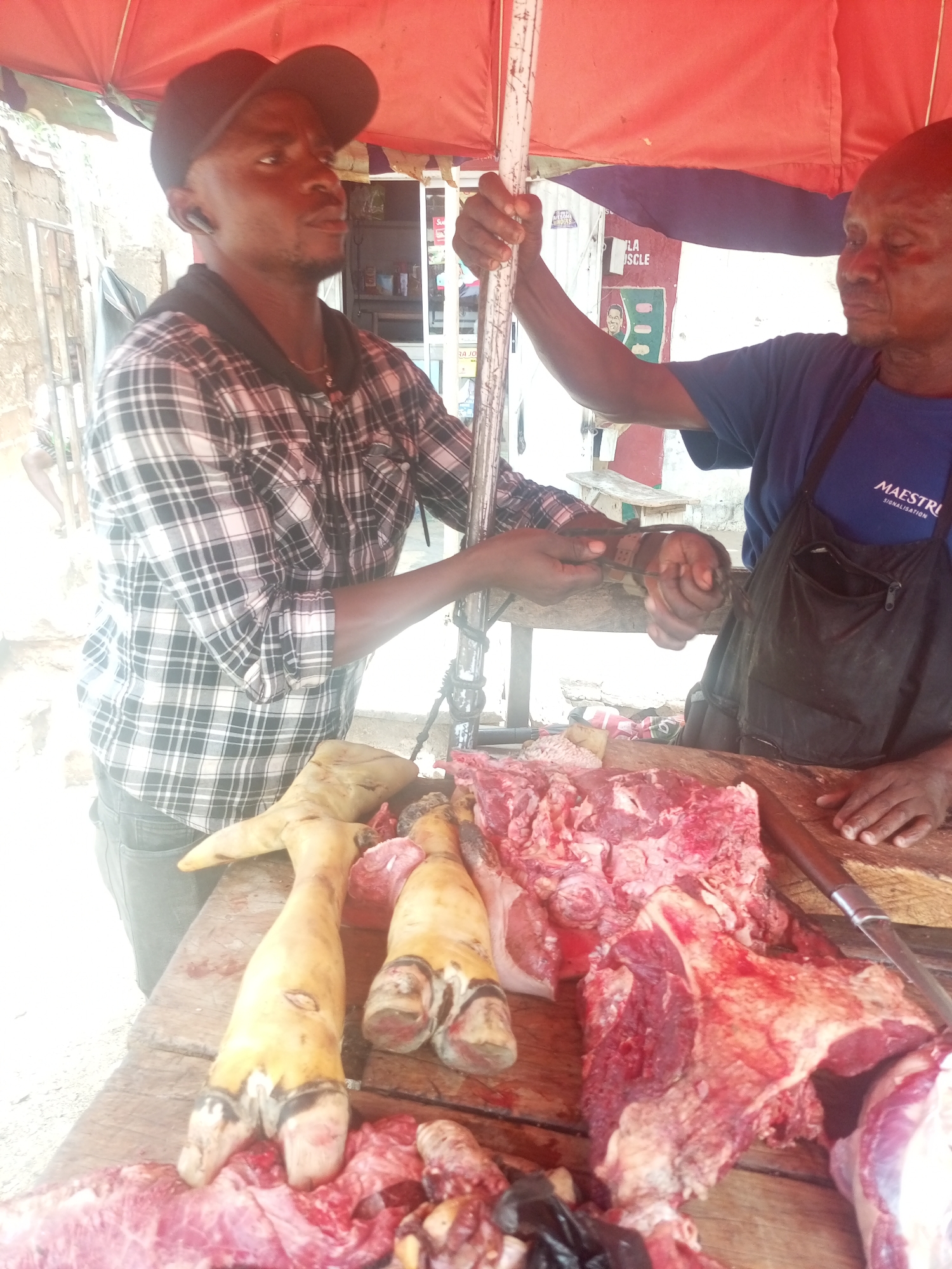 In Sabon Tasha, Nigeria, two men stand behind a wooden table under a red awning. One man wears a black cap and a black and white plaid shirt. He holds onto a white pole. The other man wears a blue t-shirt with "MAESTRI" printed on it and a black apron. He is handing a black object, possibly a wallet or small bag, to the first man. The table is covered in raw meat, including what appears to be animal hooves and cuts of red meat. A large knife is visible on the table. A building is partially visible in the background.