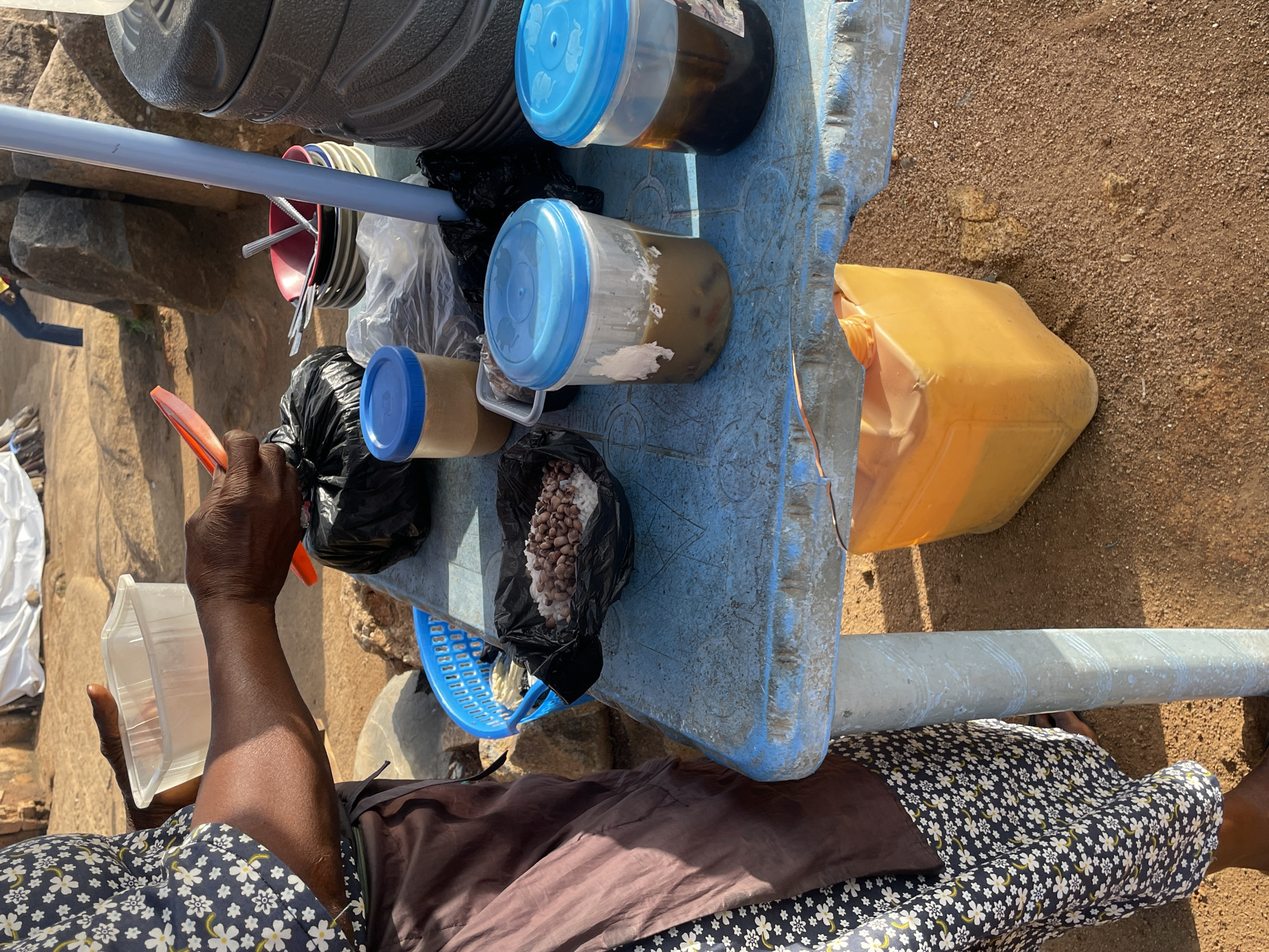 The image depicts an outdoor scene, likely in Jos, Nigeria, featuring a food vendor preparing a meal. A person, wearing a patterned blouse and a brown apron over a floral skirt, is holding a clear plastic container and a red serving utensil. In front of them, on a blue plastic table, are several items. These include a black plastic bag containing rice and beans, a blue-lidded plastic jar with a tan substance, another blue-lidded plastic jar with a brown liquid, and a stack of bowls. A large gray cooler is positioned to the right of the table. A blue plastic basket is visible on the table, and an orange plastic container is situated beneath the table. The ground is covered in reddish-brown soil and small stones, and large rocks are visible in the background, suggesting a semi-arid or rocky terrain. The lighting indicates it is daytime.